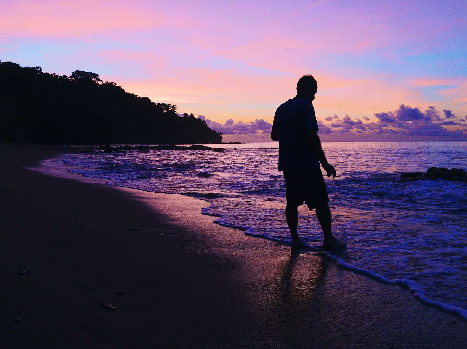 A man smokes a cigarette while getting his feet wet on a beach. Sunset. 