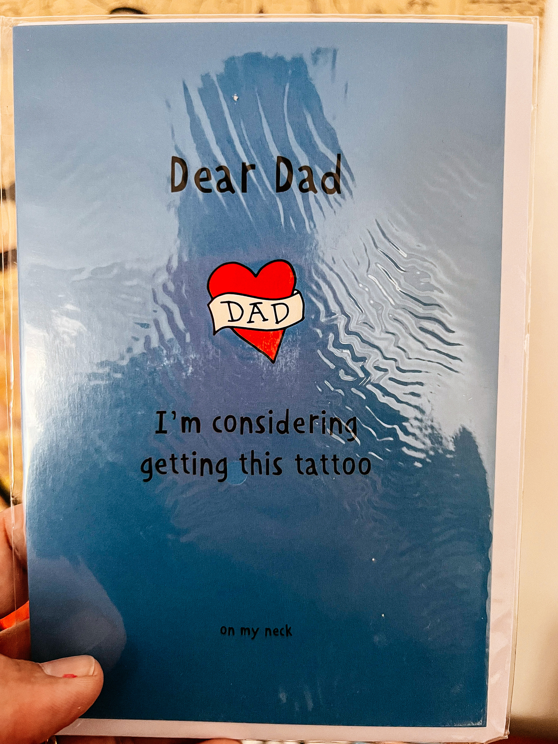 a postcard with a tattoo design of a heart with the word “dad” over it, and “Dear dad, I’m considering getting this tattoo… on my neck”. 