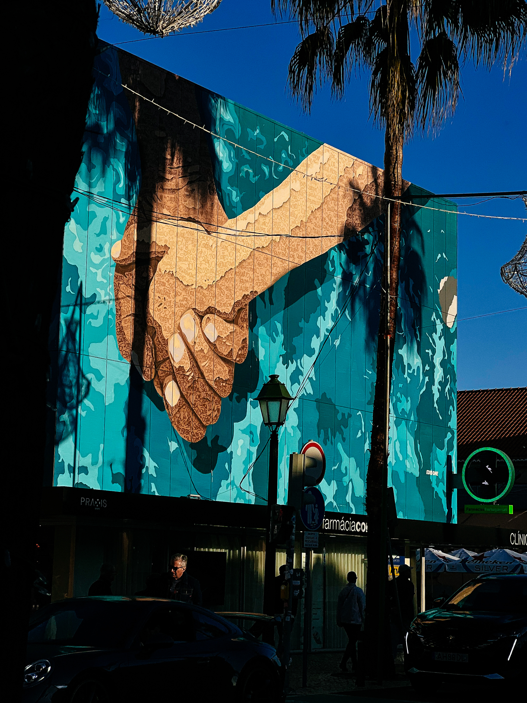 A mural on a building featuring a large hand holding a smaller hand, amid abstract blue patterns. In the foreground, shadowy figures of people, cars, a streetlight, and palm trees against a bright sky.