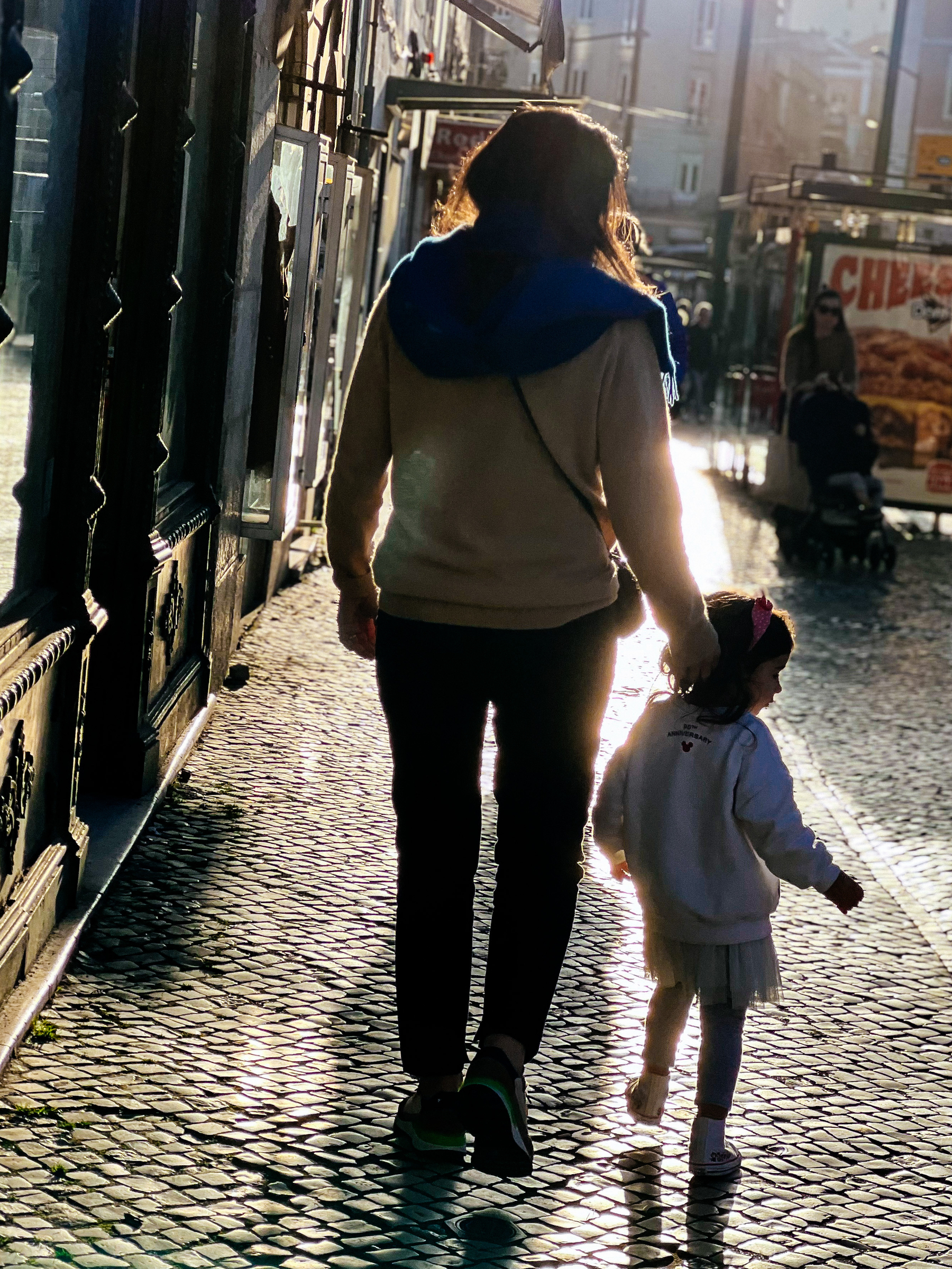 A mom and daughter couple walk down the street in a city environment, their backs to us. The girl is looking to her right, and the mom gently steers her in the right direction 