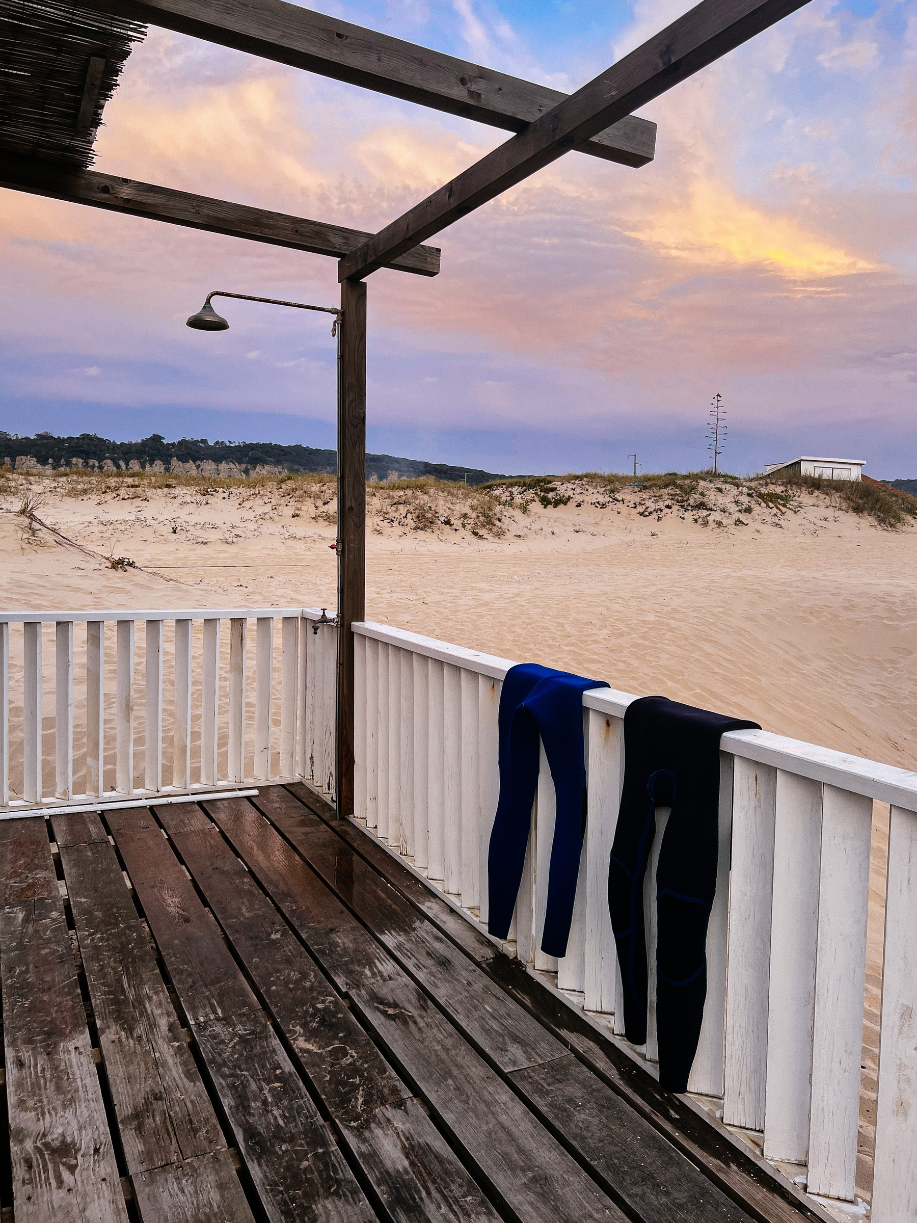 Two wetsuits hanging from a fence. A shower on the left side, and sand on the background. 