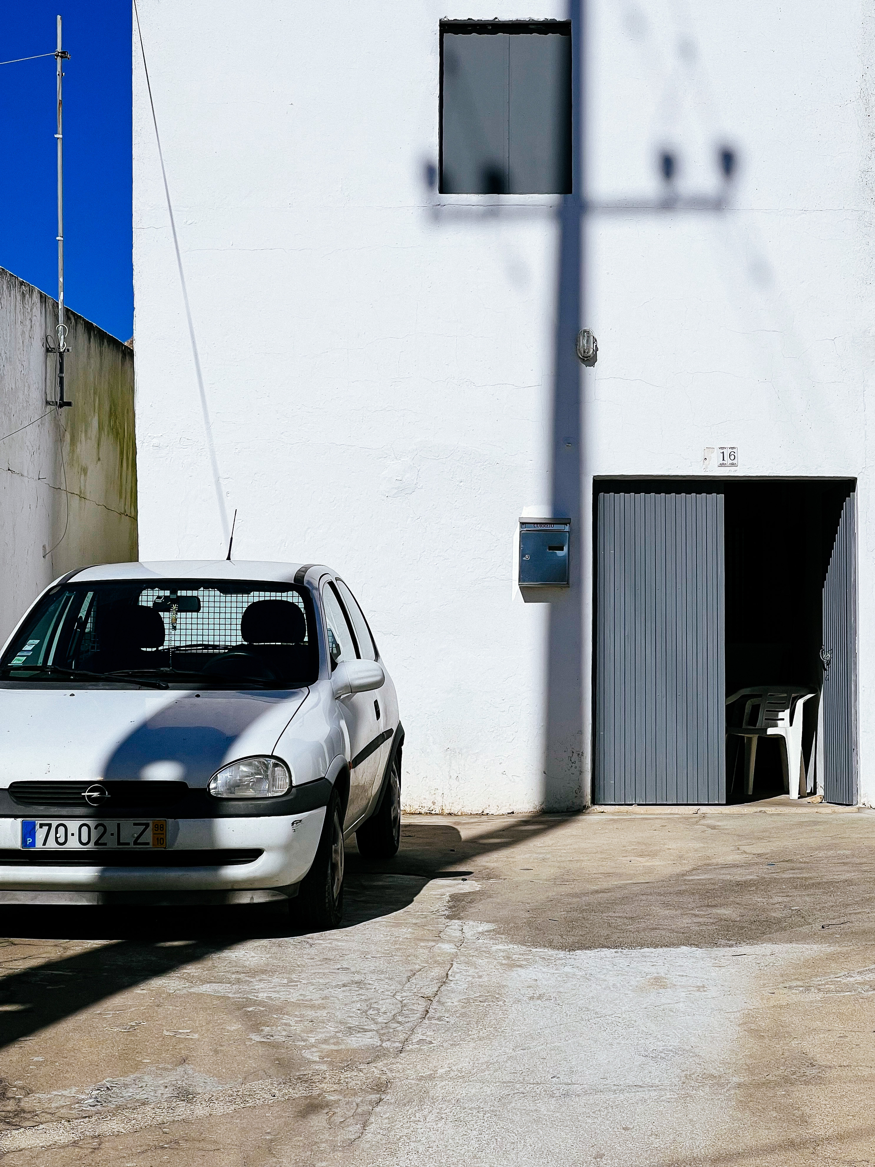 A white car parked in front of a white building, an ugly one. Shadows falling on both the car and the building. The whole scene is white.