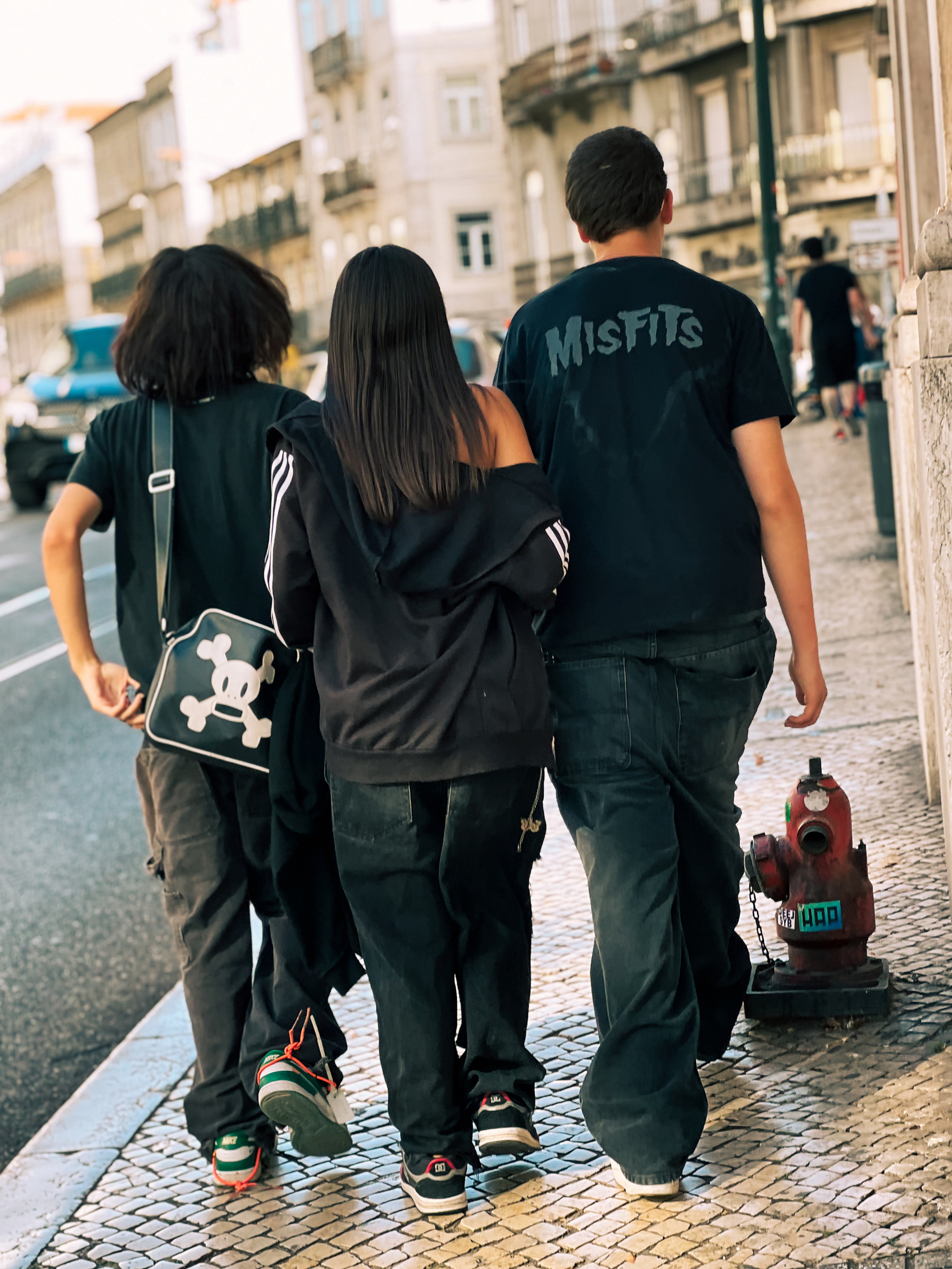 Three goth enthusiasts walk away from us. This-shirt says Misfits. 