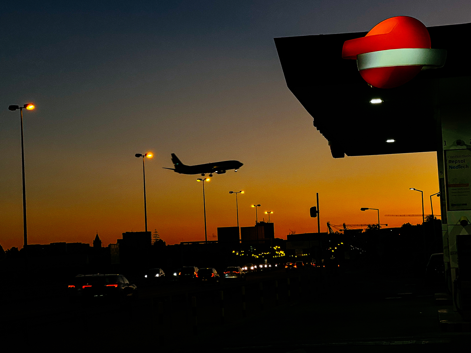 An airplane approaching the airport, flying over a road, next to a gas station. During sunset. 