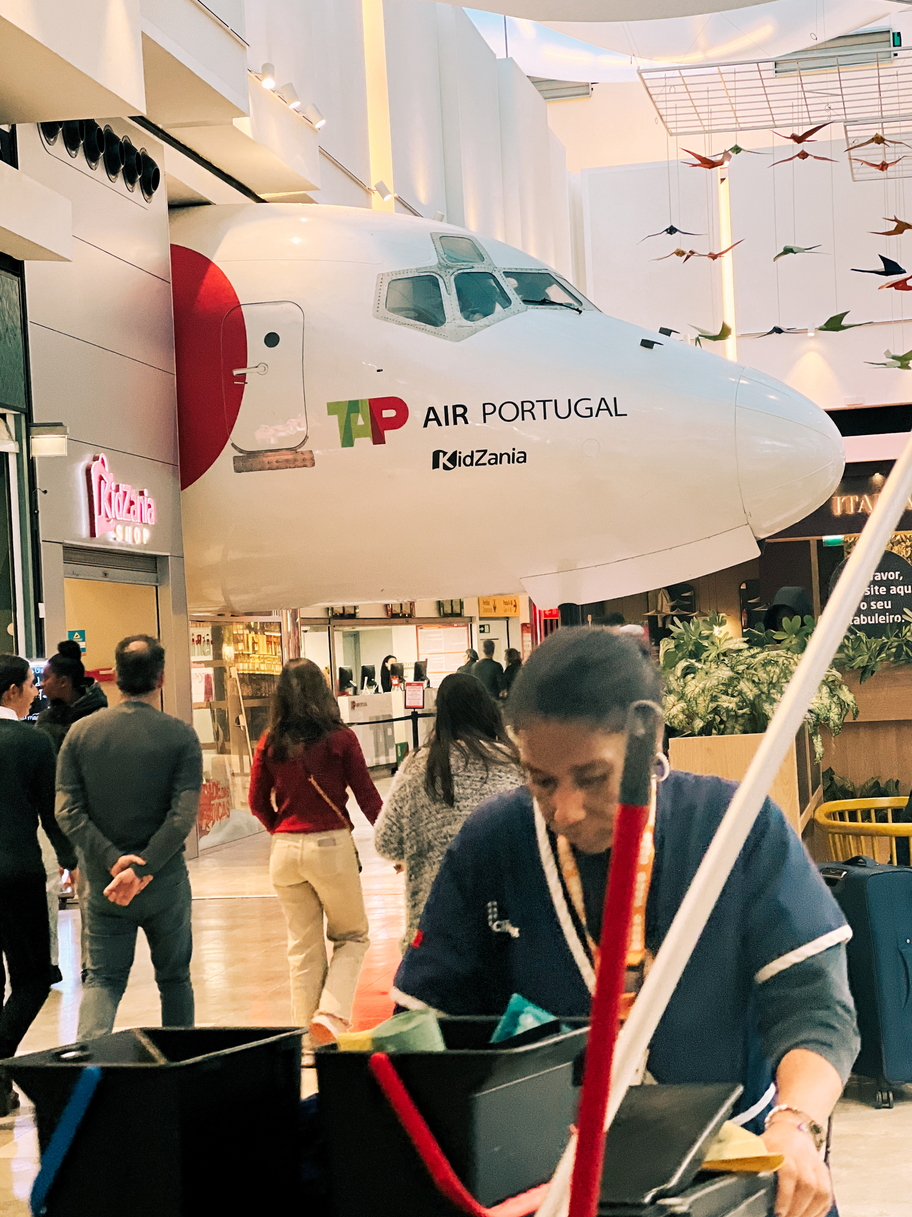 A shopping center with a large model of a TAP Air Portugal aircraft prominently displayed. Below, people are walking around, and a person is in the foreground cleaning with a cart of supplies.