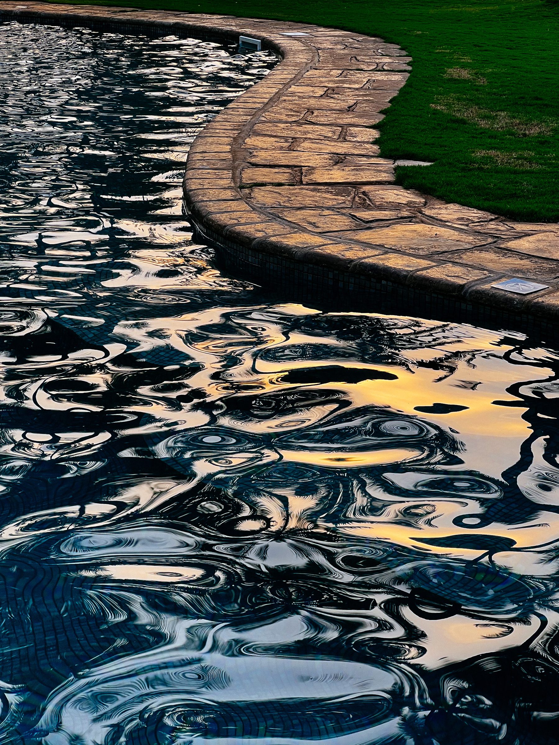 The sun is reflected on water in a swimming pool. We can still see some grass on the side of the pool. 