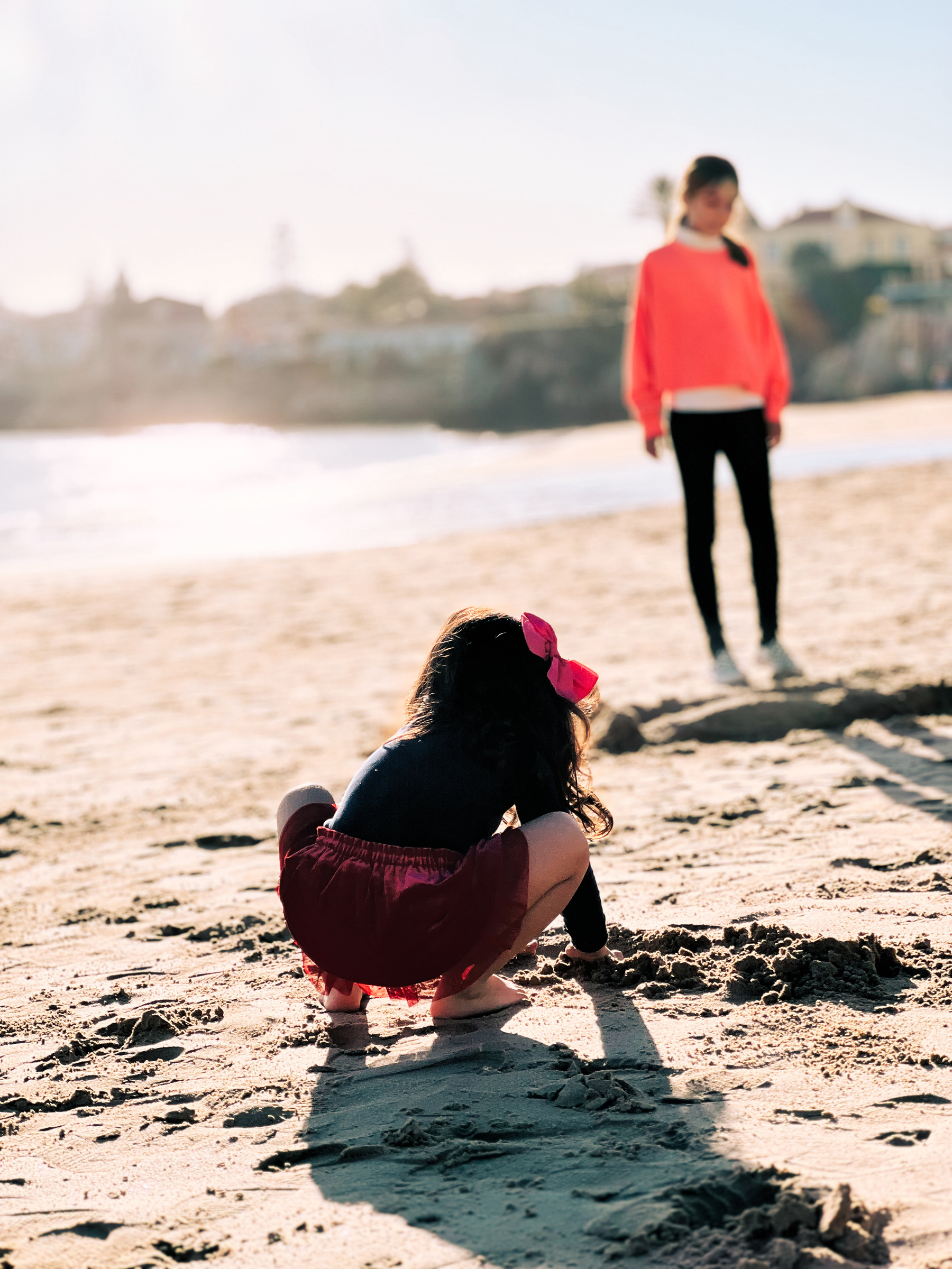 A young girl in a red skirt and black top crouches on a sandy beach, playing in the sand, with a girl standing in the background looking on.