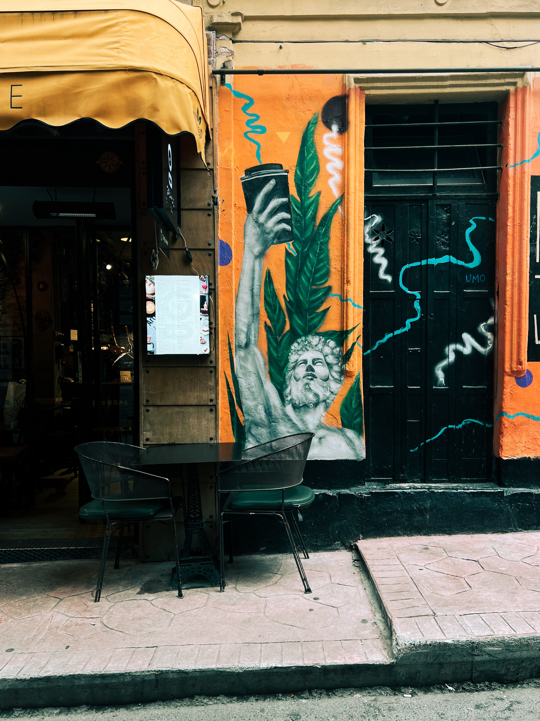 A colorful street mural depicting a figure holding a coffee cup alongside green foliage, with a bistro table and two chairs set up in front of it on the sidewalk.