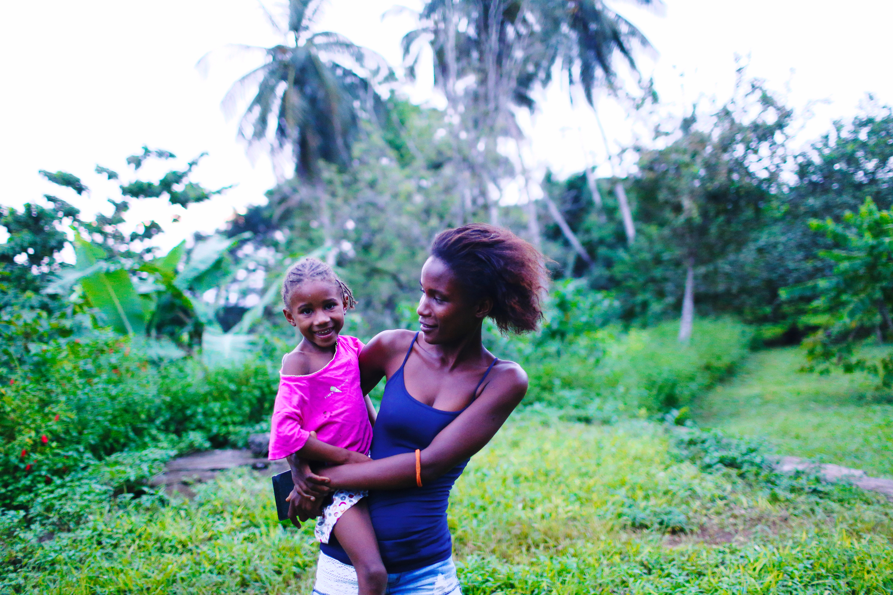 A girl holds a baby, both smiling. They’re standing in a tropical forest. 