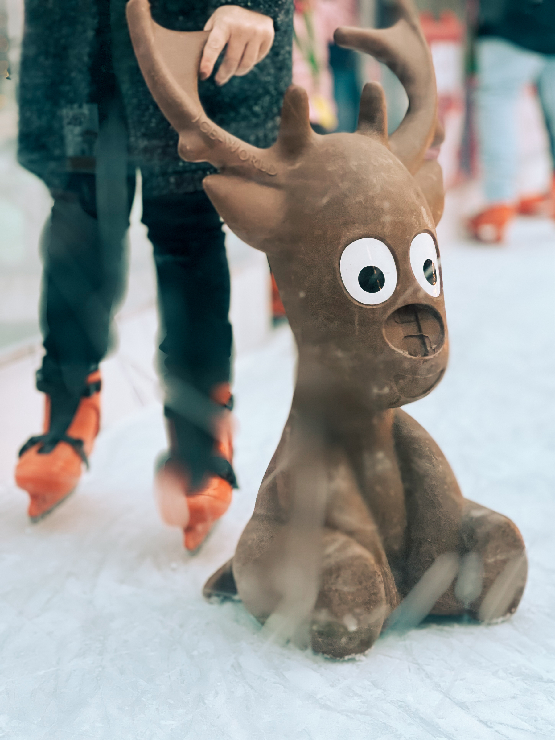 A plastic reindeer skating aid on an ice rink with a blurry figure of a person in ice skates standing behind it.