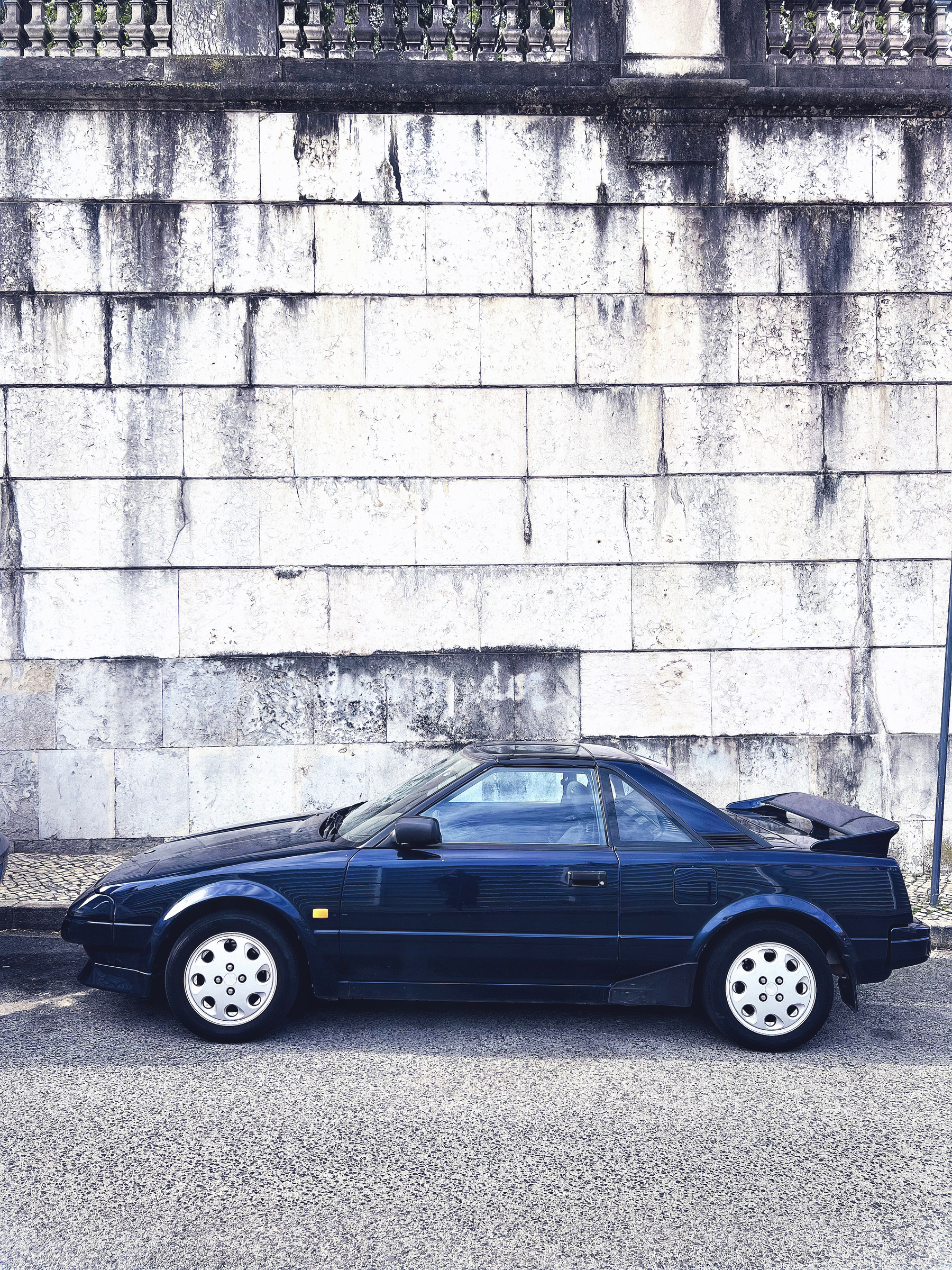 a dark blue car, unknown brand, against a white stone wall