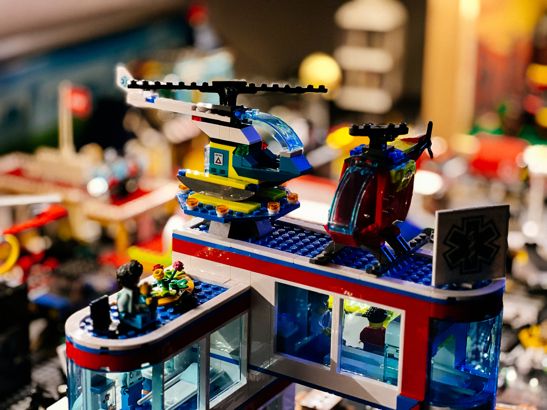 A detailed LEGO set depicts a hospital scene with a helicopter landing pad on the roof where two colorful LEGO helicopters are stationed. Minifigures are placed around, suggesting activity and storytelling within the setup.