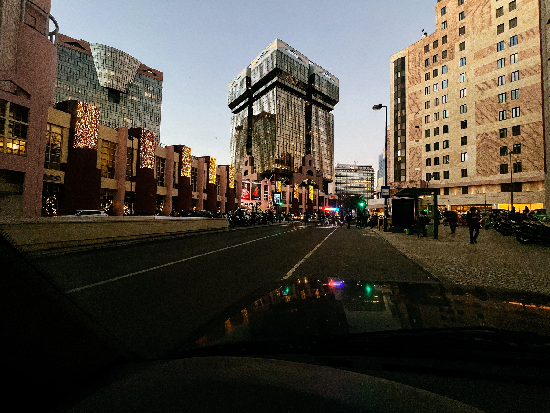 A city street view at dusk with modern buildings, some decorated with lights, as seen from a car&rsquo;s perspective. There are pedestrians, motorcycles parked on the sidewalk, and signs of urban activity.