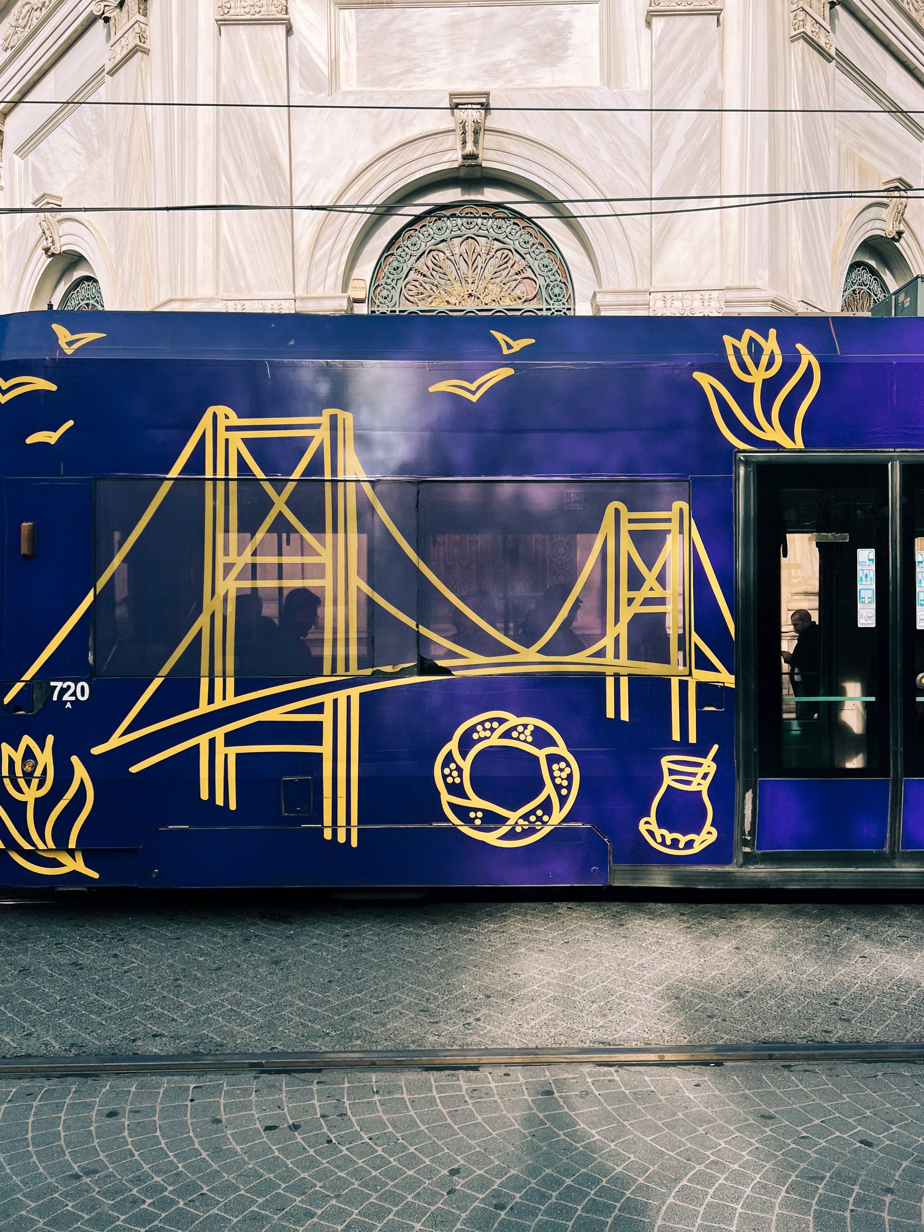 A blue streetcar with decorative yellow line graphics, including a bridge and traditional symbols like a pretzel and pitcher, parked in front of a building with detailed stonework and an ornate window.