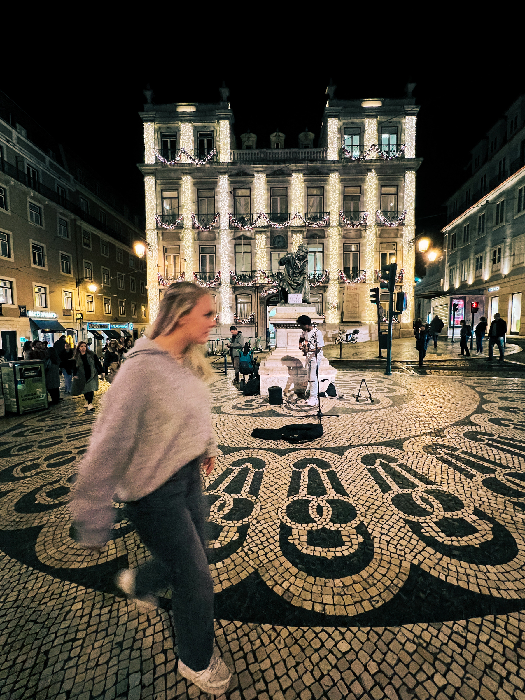A busy street scene at night with a person walking in the foreground, a street performer in the middle ground, and an ornately decorated building covered in festive lights in the background. The ground features a traditional black and white mosaic pavement.