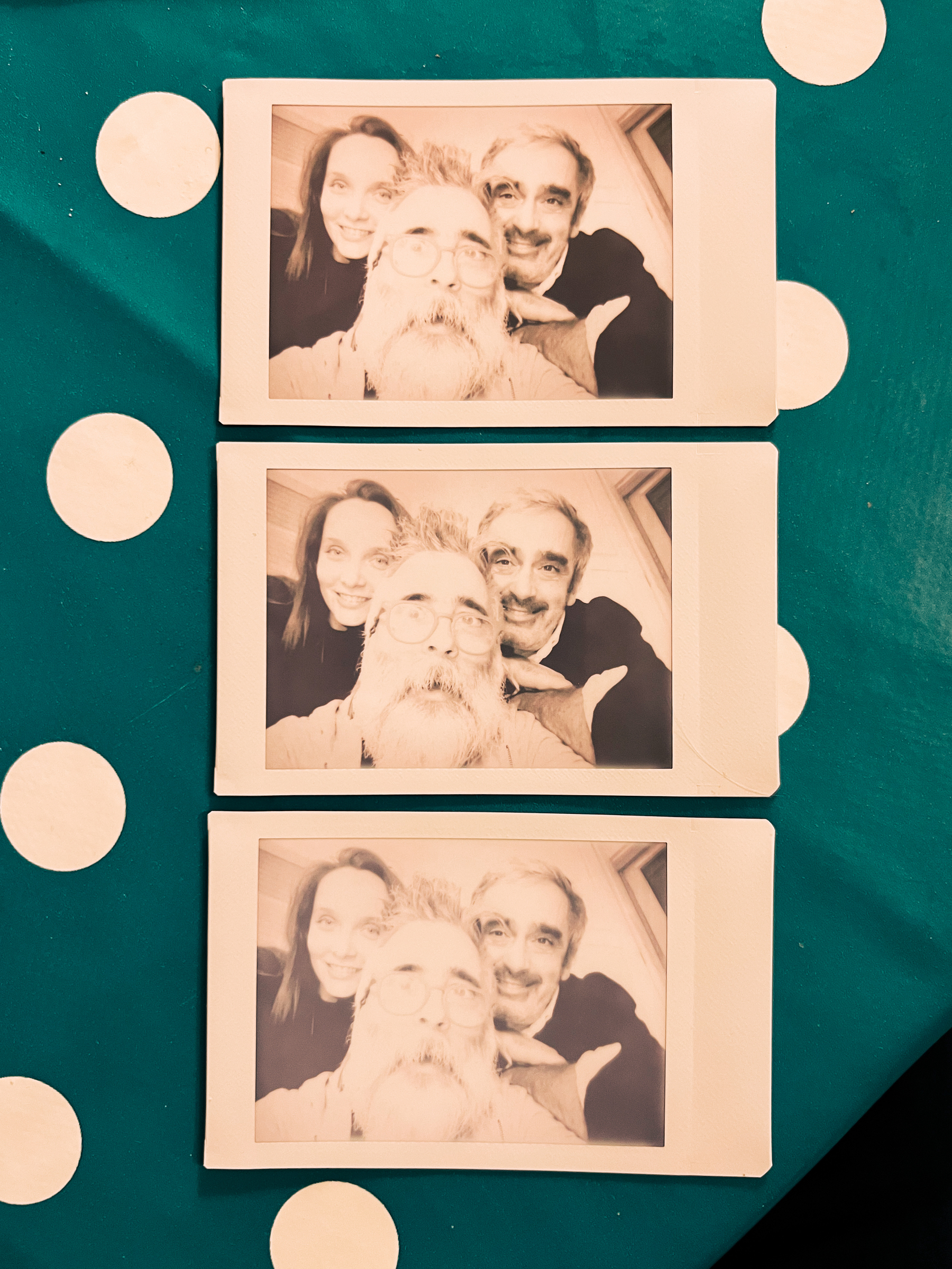 Three Polaroid photos positioned on a green surface with white dots, each showing the same three smiling people posing closely together for a selfie.