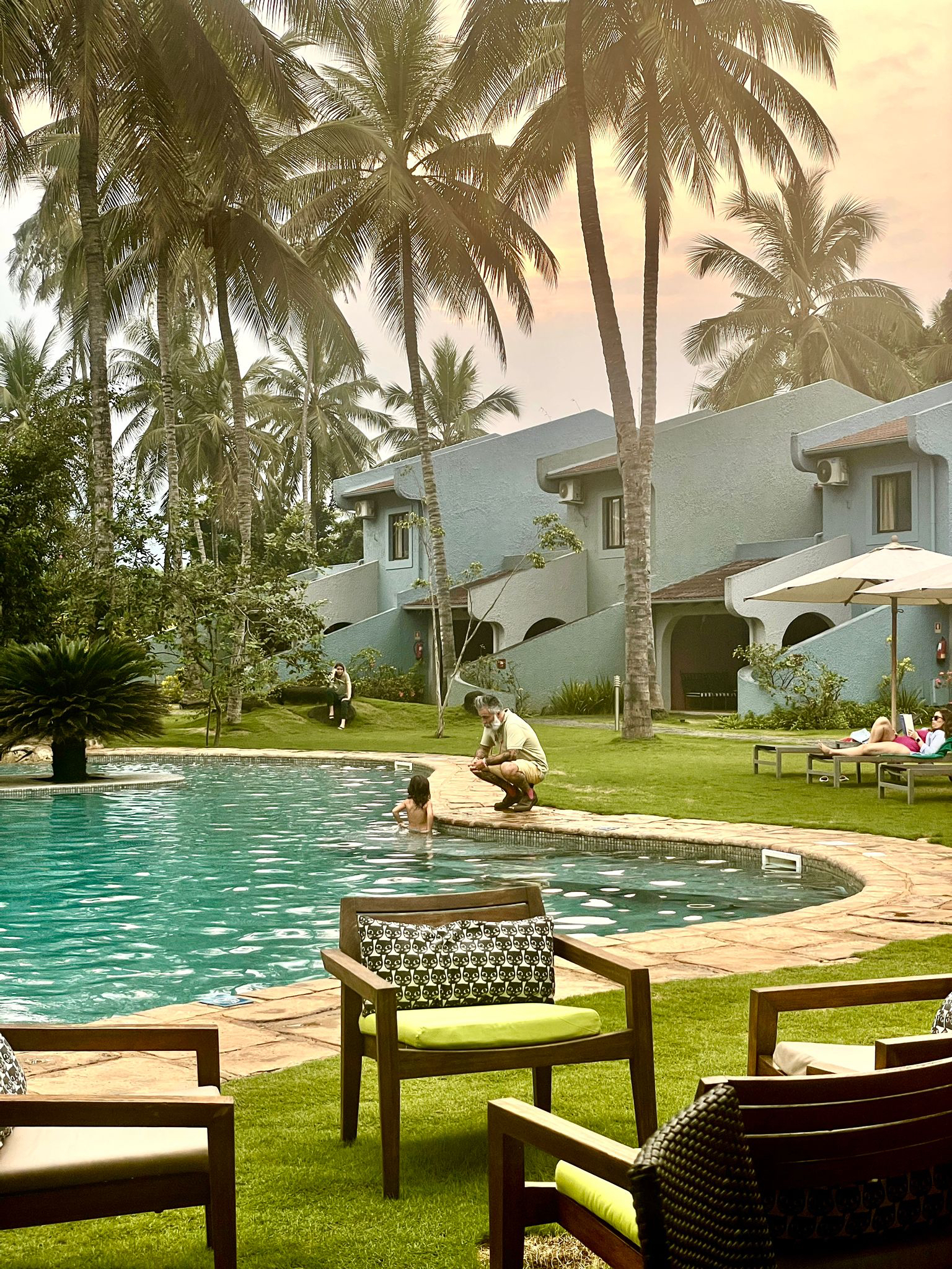 A swimming pool, with a toddler in it, talking to her father, who’s standing outside. There are chairs in the foreground, and palm trees all around. 
