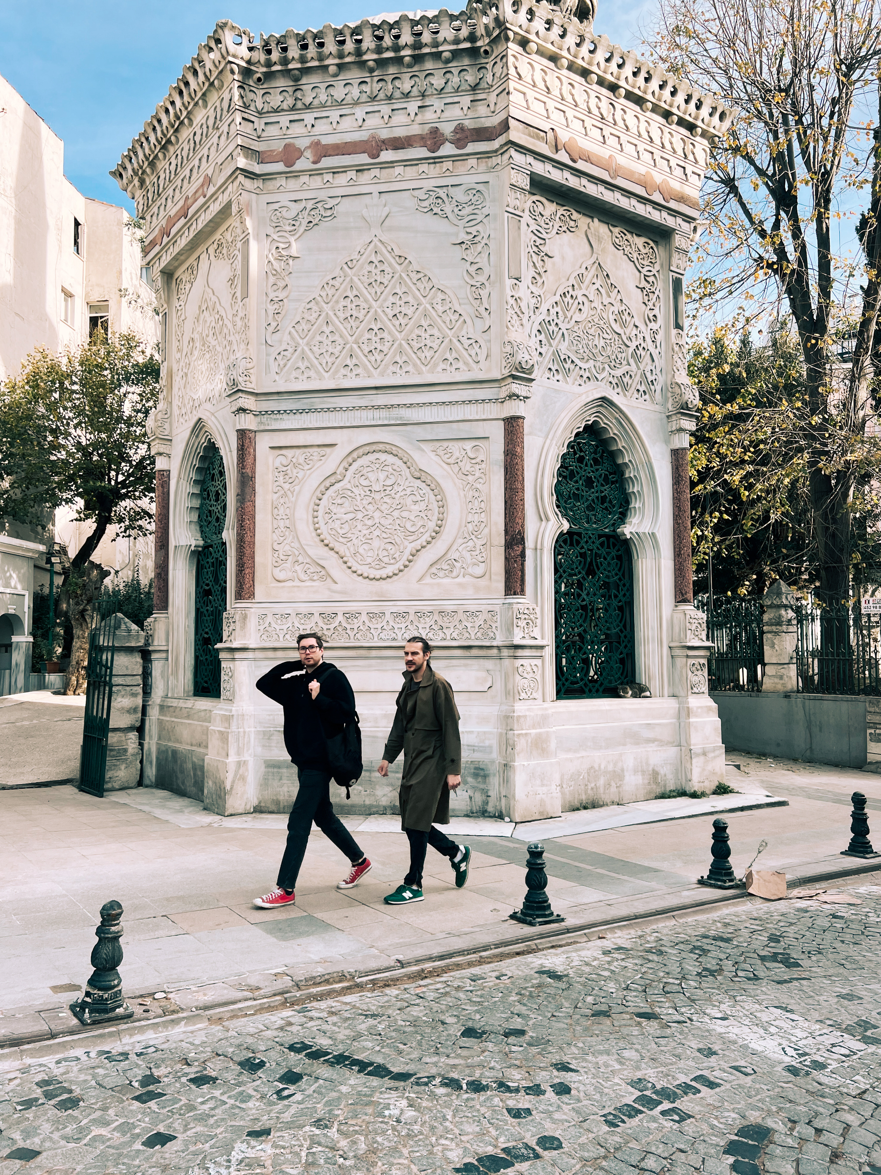 Two men walking past an ornate historic building with intricate patterns and a pointed archway.