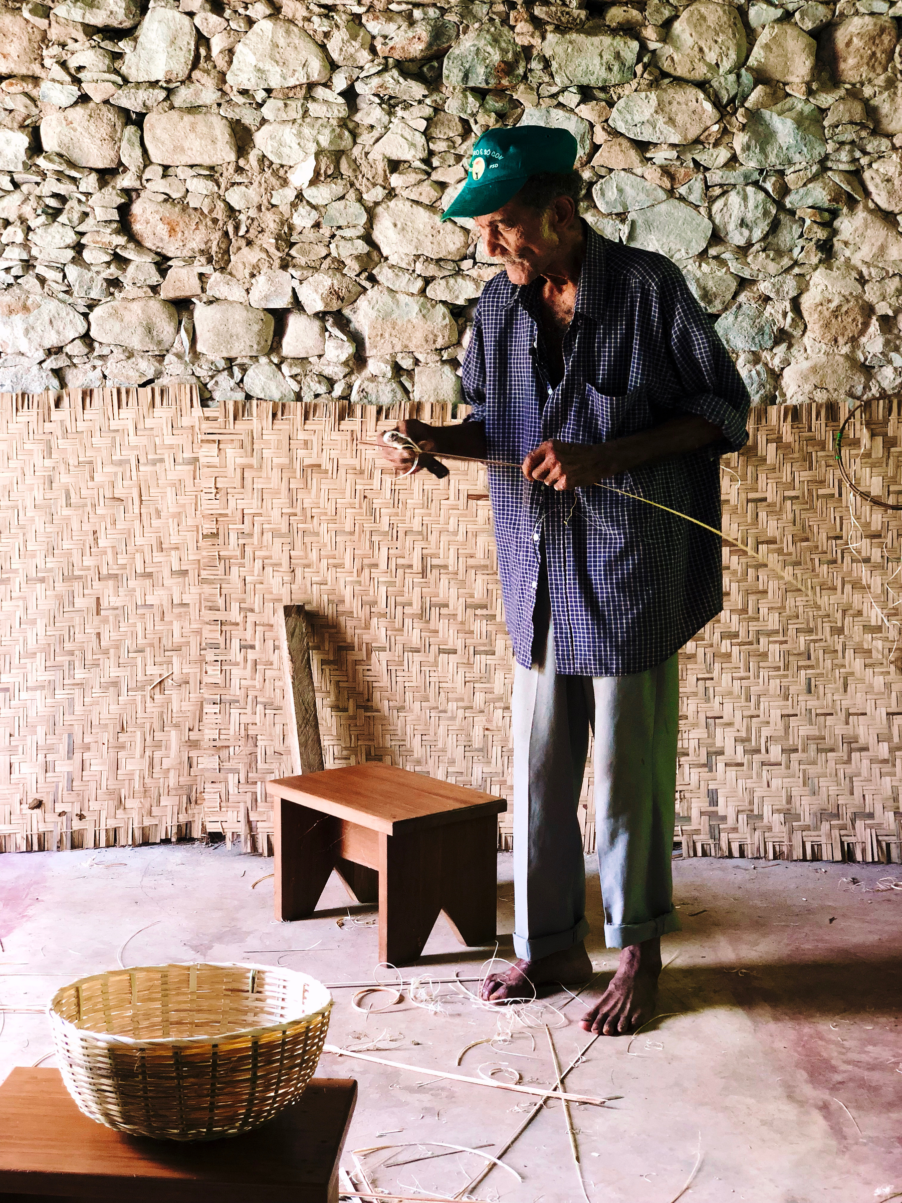 A gentleman prepares to weave. He’s standing next to a tiny stool, and a basket is seen in the foreground. 