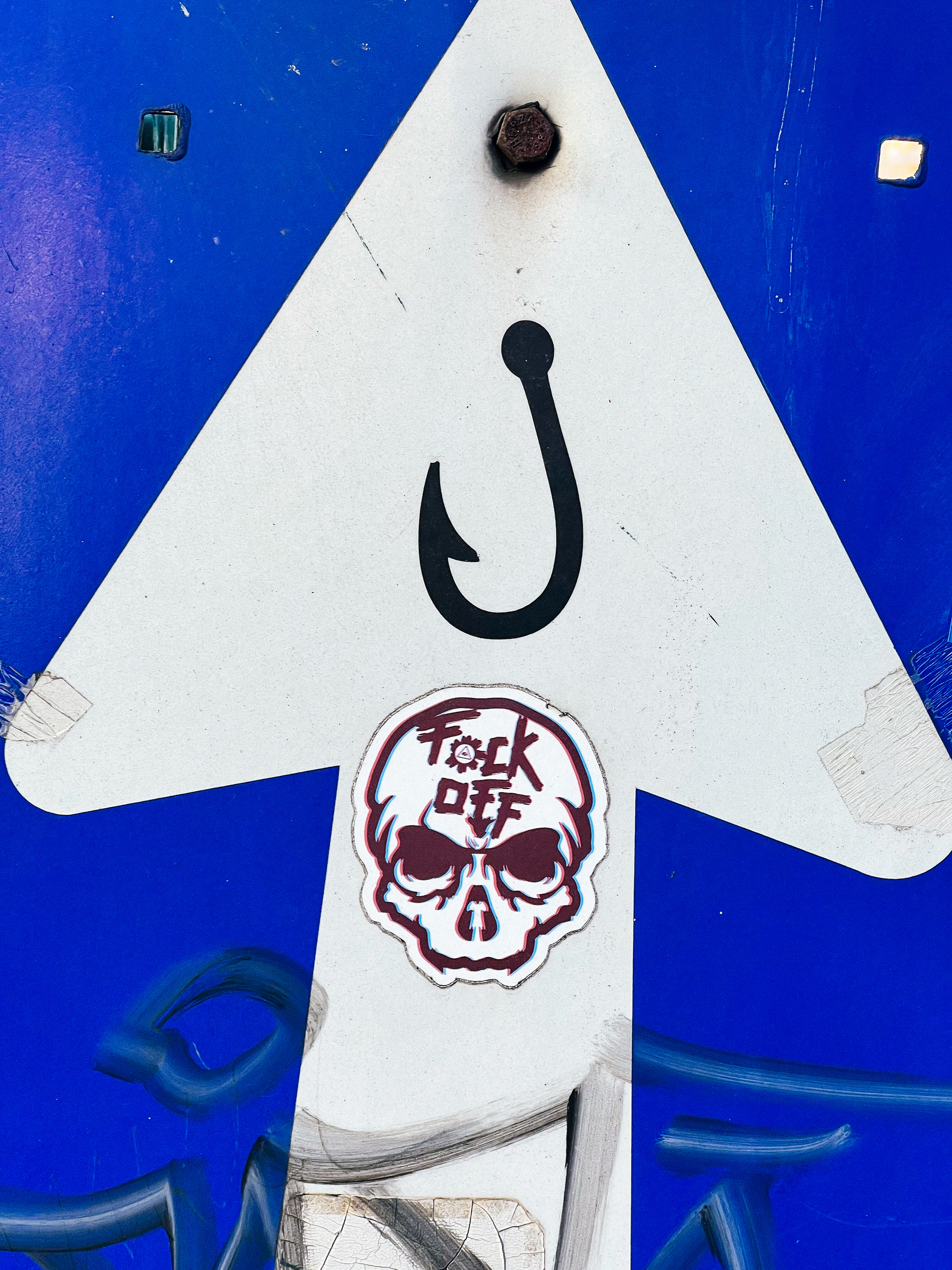 A skull with “Fock Off” written on it, and a fishing hook. Both stickers on a traffic sign. 