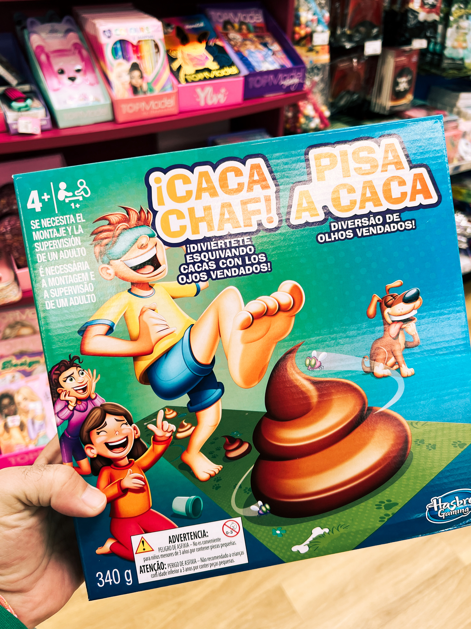 A person&rsquo;s hand holding a board game box named &ldquo;¡CACA CHAF!&rdquo; with animated images of children and a dog, and a comically large pile of poop. The game involves wearing a blindfold and avoiding stepping in fake poop.