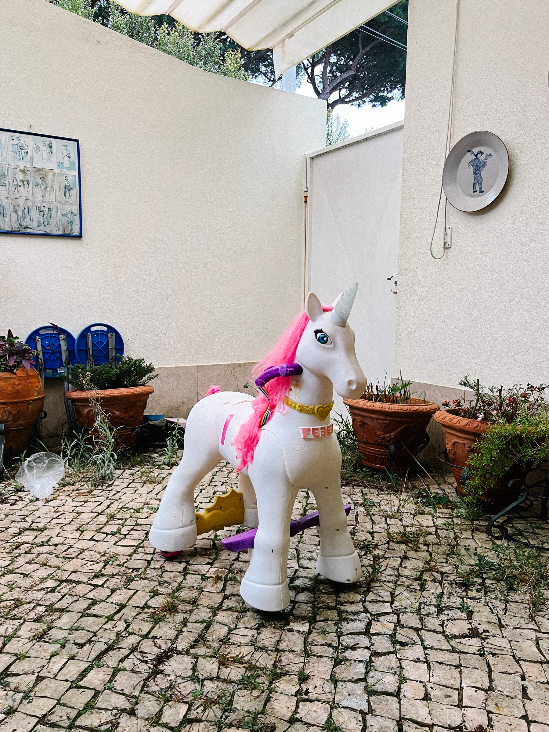 A child&rsquo;s ride-on unicorn toy with a pink mane in a courtyard with cobblestone flooring, surrounded by potted plants, a white awning, and house walls with decorative items.