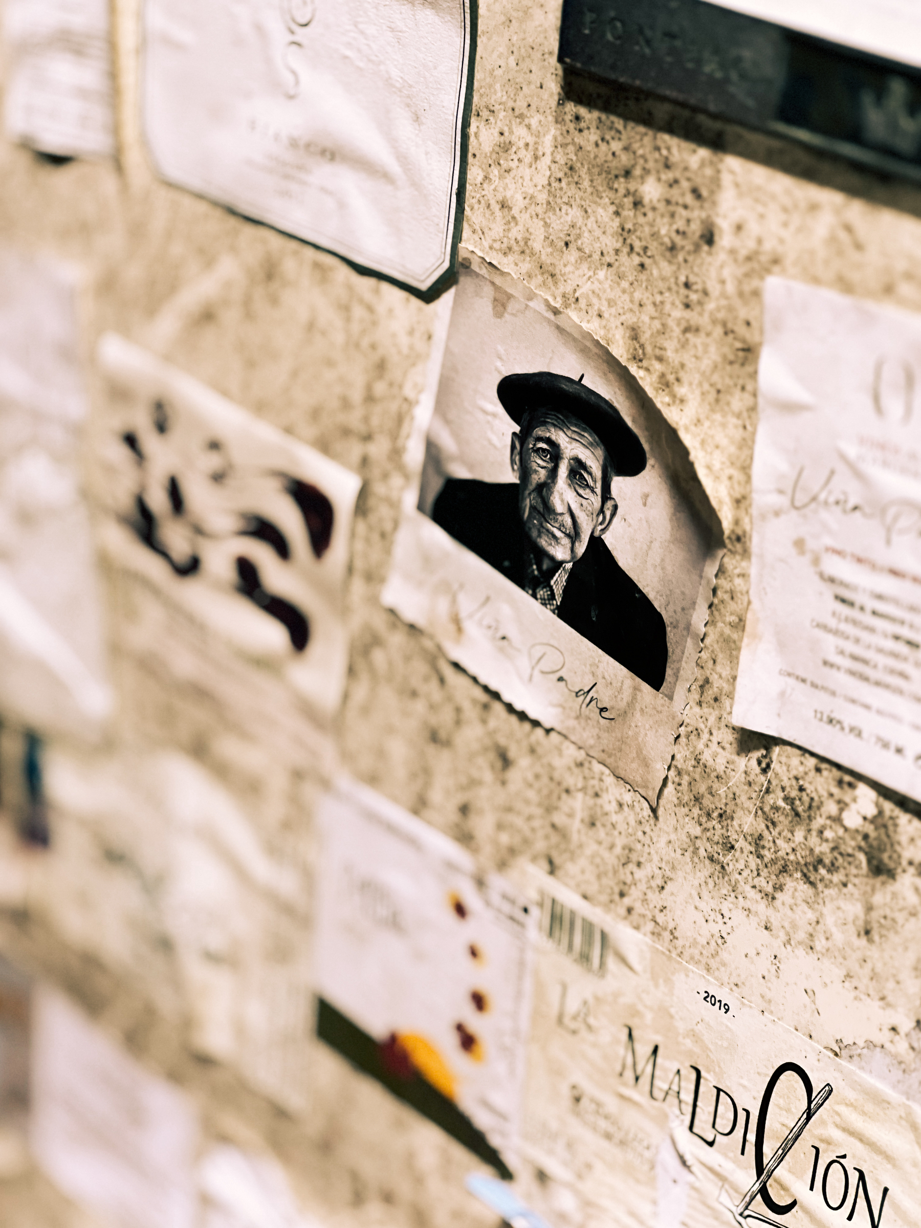 A tilted and close-up view of a wall covered with various notes and cards, with a photo of an elderly man wearing a beret centered in the image, surrounded by other papers.