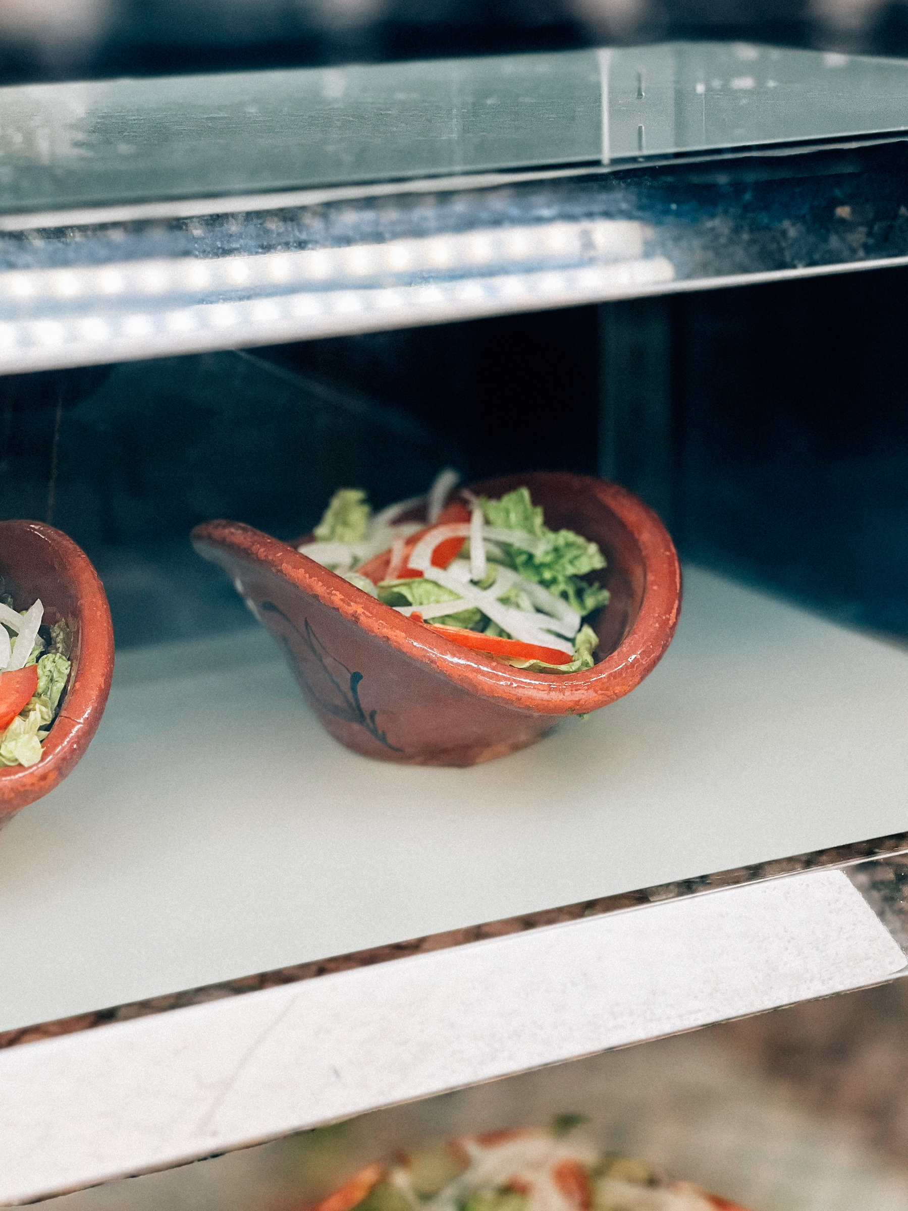 A clay bowl containing fresh salad, consisting of lettuce, onion, and tomato, displayed in a refrigerated case.
