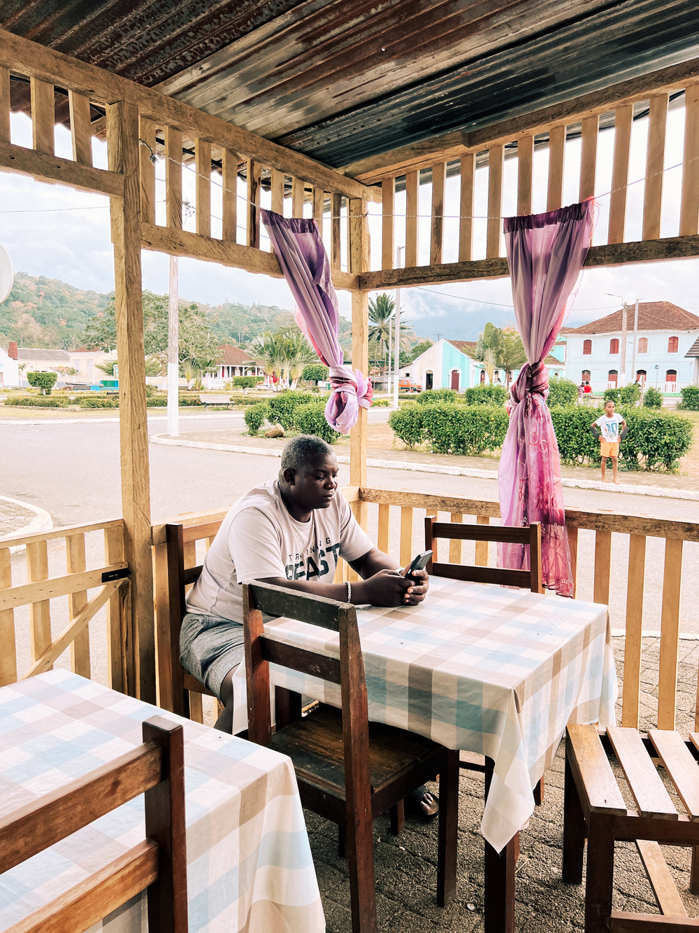 A person sits alone at a wooden table in an open-air structure, looking at a mobile phone. The table is covered with a checkered tablecloth. Purple curtains tied to the wooden posts frame the open space.