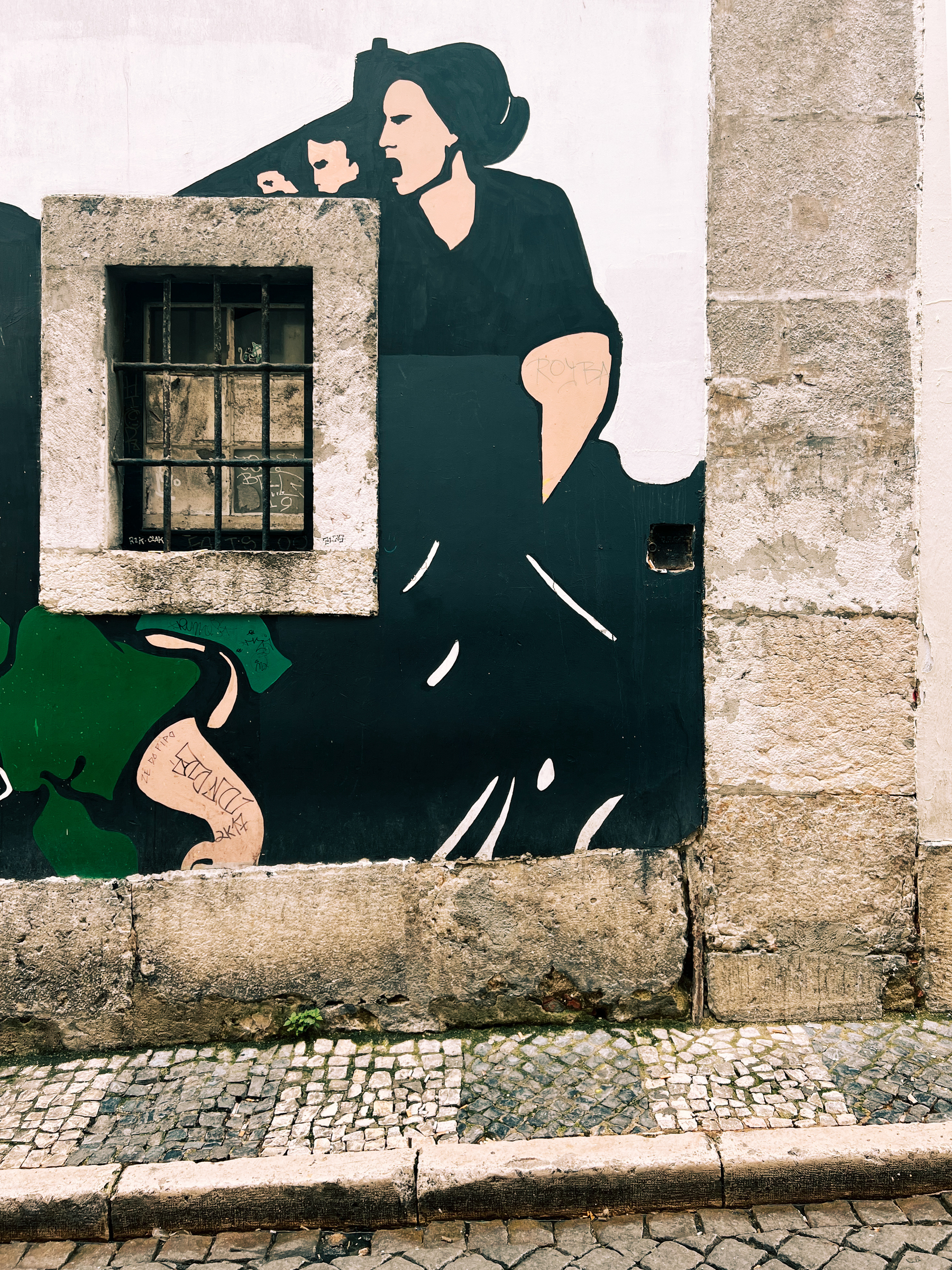 A mural on a building wall featuring a stylized black and white figure of a woman, integrated with a real window that acts as part of the art piece. The cobblestone pavement and step in front of the wall add texture to the scene.