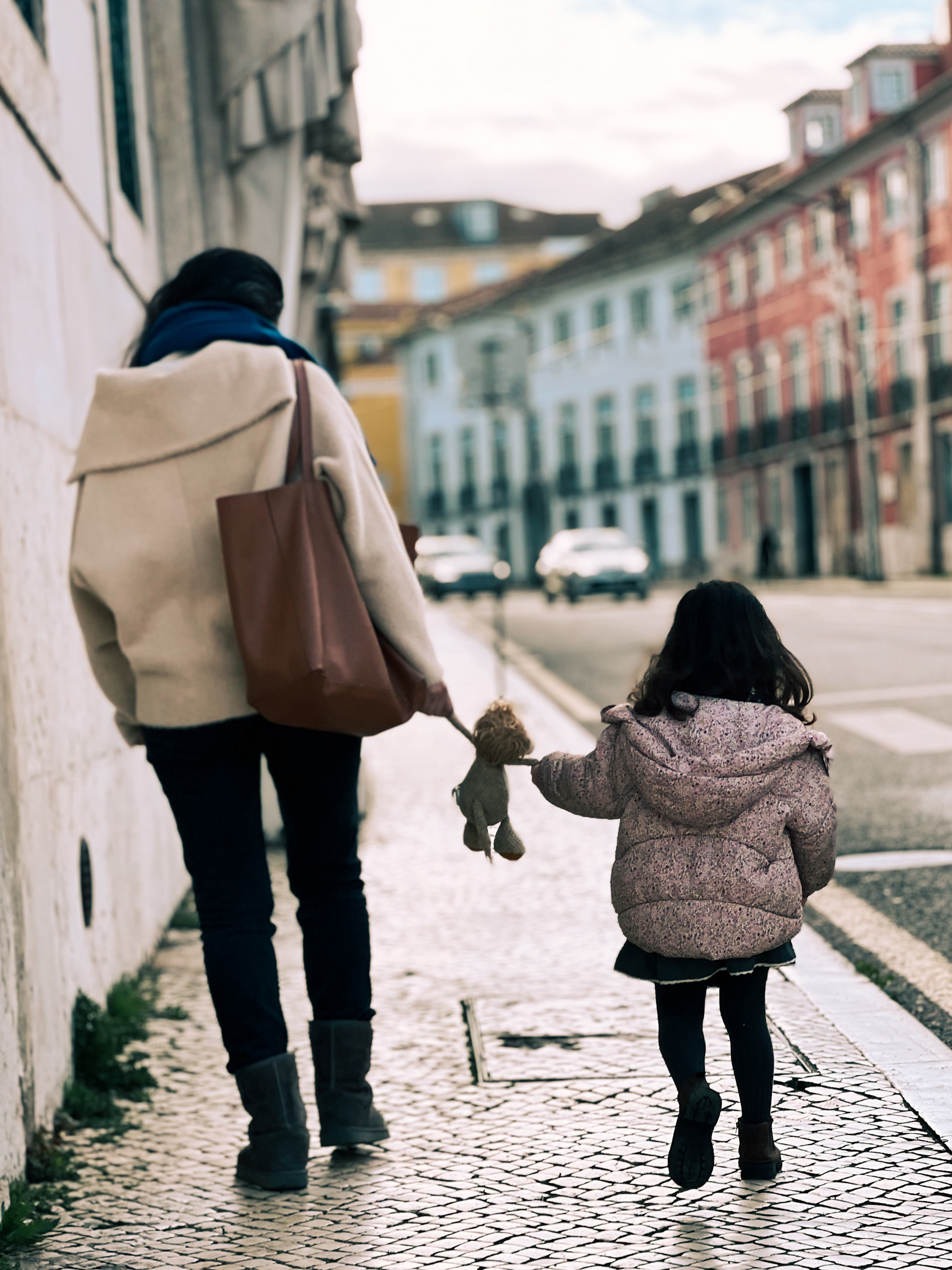 Adult and child walking hand in hand down a cobblestone street, holding a stuffed animal, with historic buildings in the background.