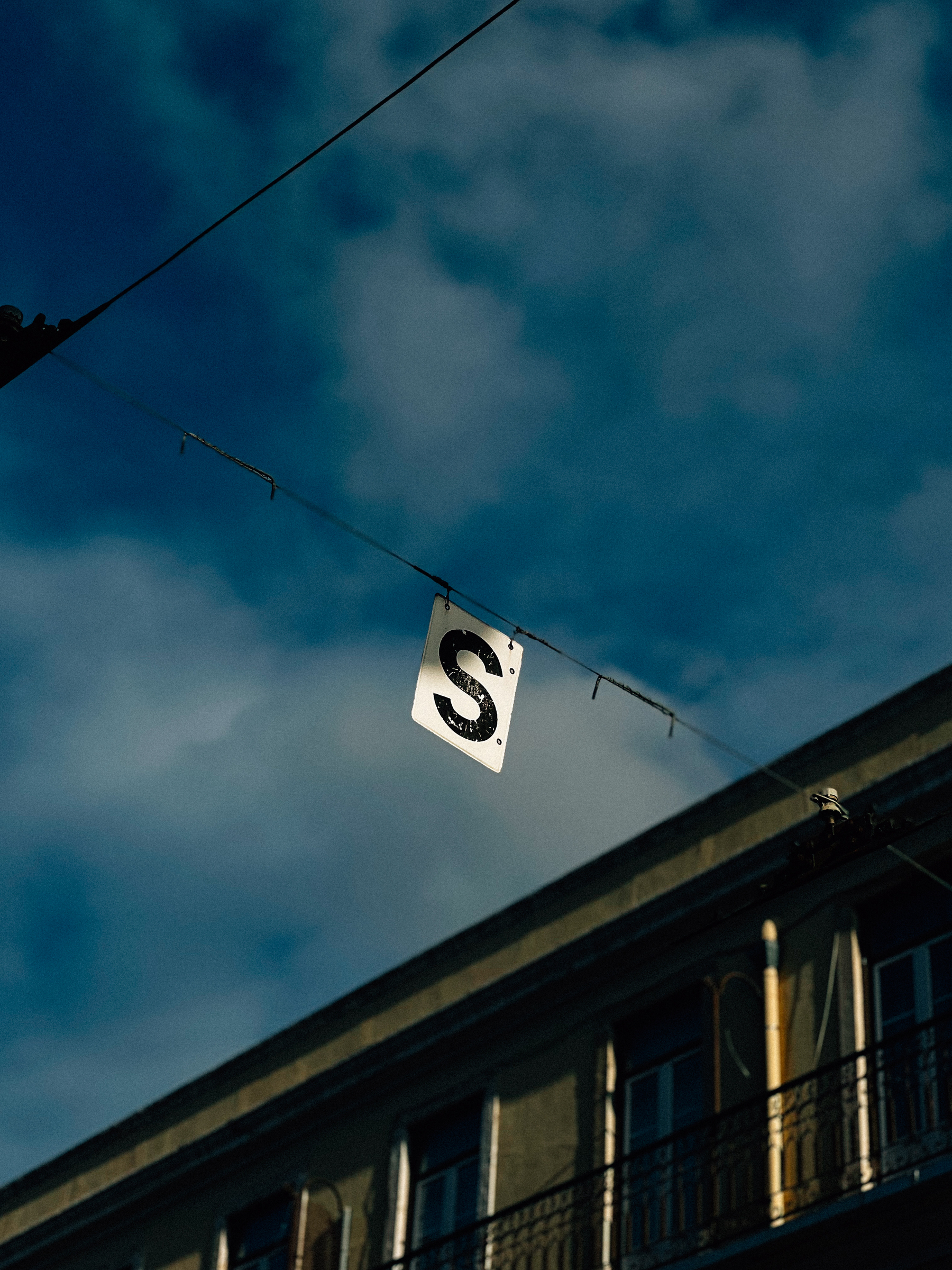 A sign with the letter &lsquo;S&rsquo; hanging from a wire against a backdrop of a blue sky and the upper facade of a building with balconies.