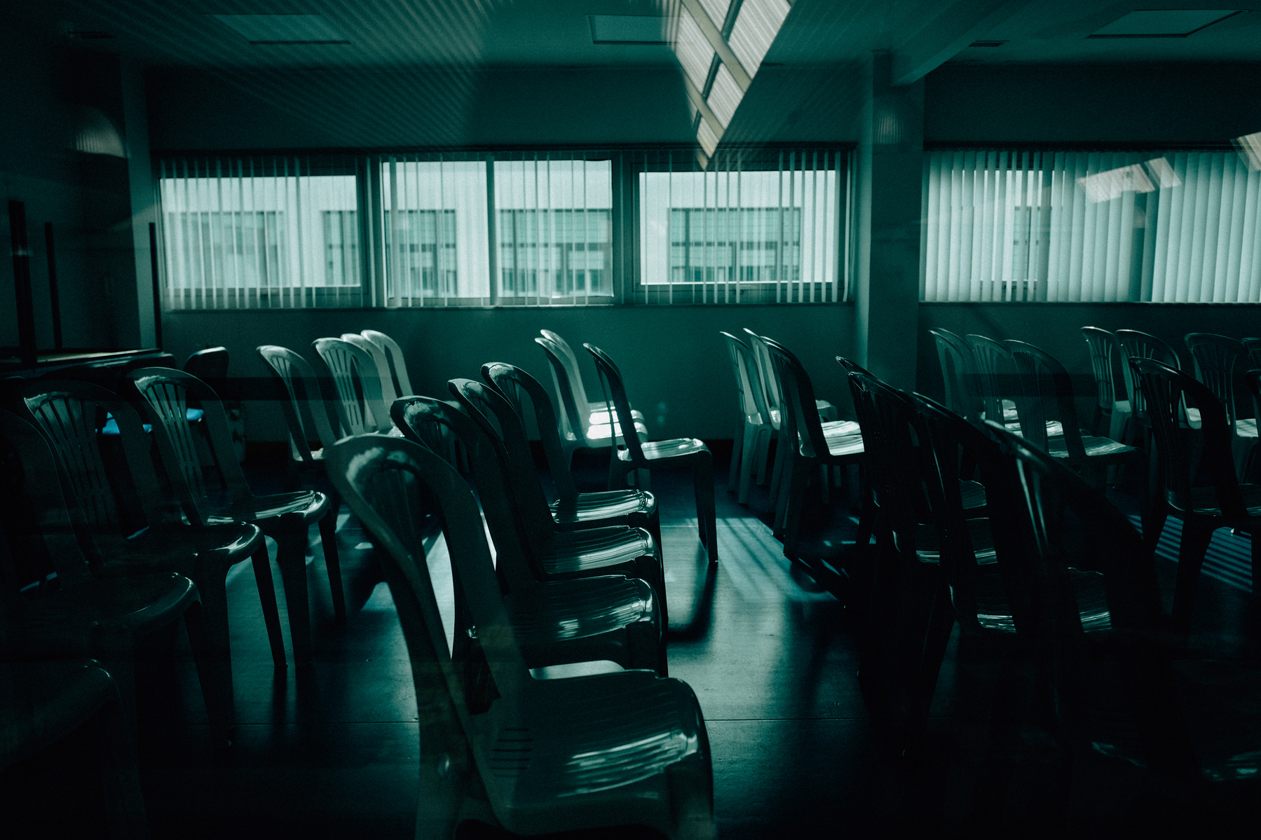 An empty room with rows of plastic chairs and tables, with sunlight casting shadows through blinds on windows and a skylight.