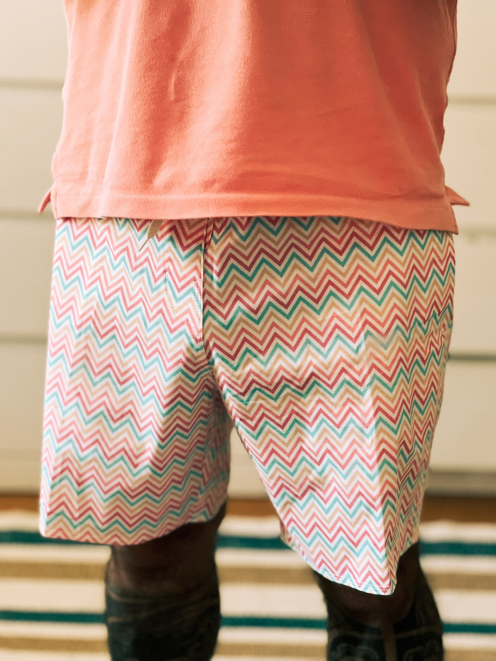 A person wearing a peach-colored shirt and multicolored zigzag-patterned shorts stands on a striped rug. The photo is centered on the person&rsquo;s lower torso and upper legs.