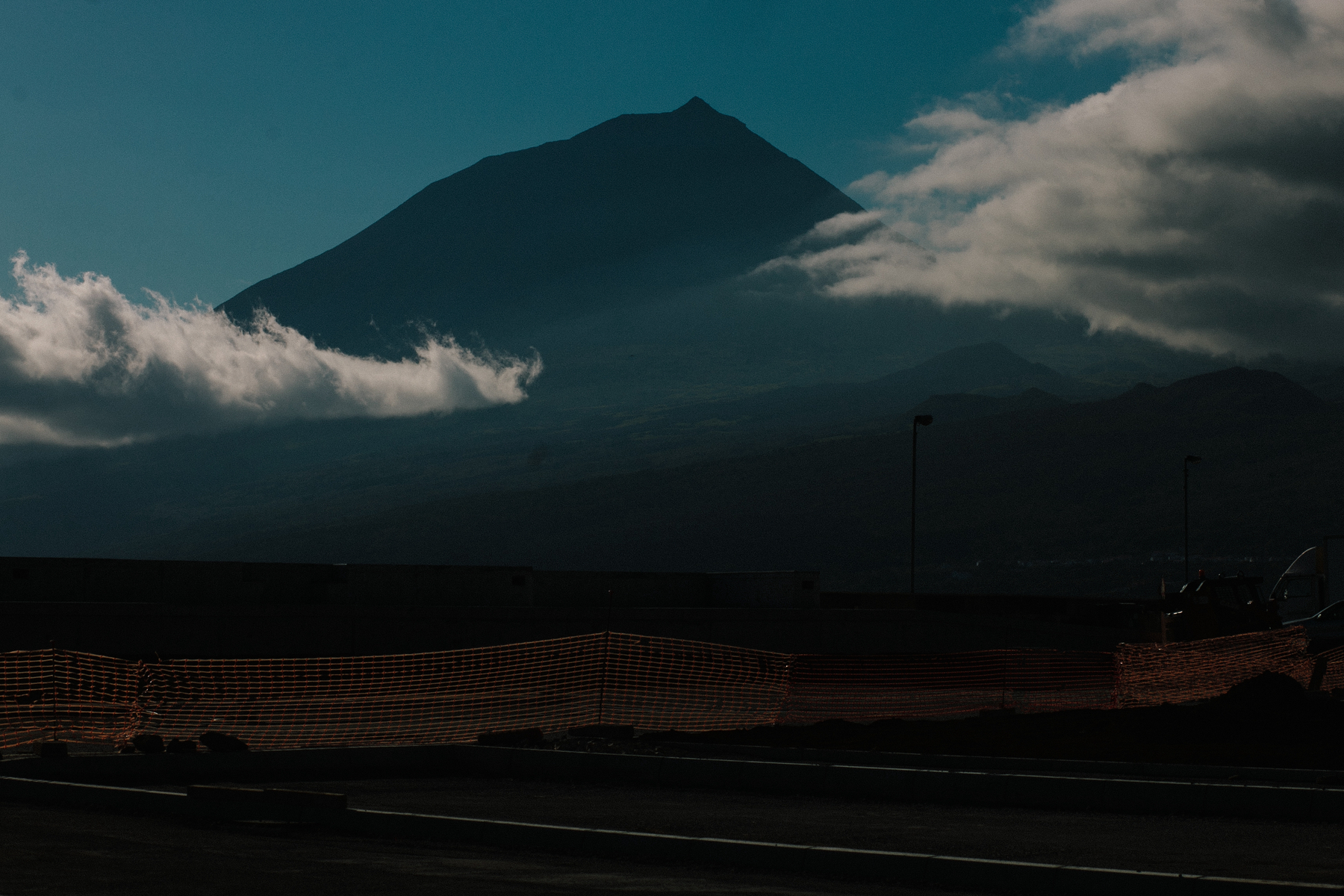 A majestic mountain looms in the distance, partially shrouded by clouds, under a blue sky. In the foreground, there is a fence and the edge of a paved area, possibly a road or parking lot. The lighting suggests either dawn or dusk. 