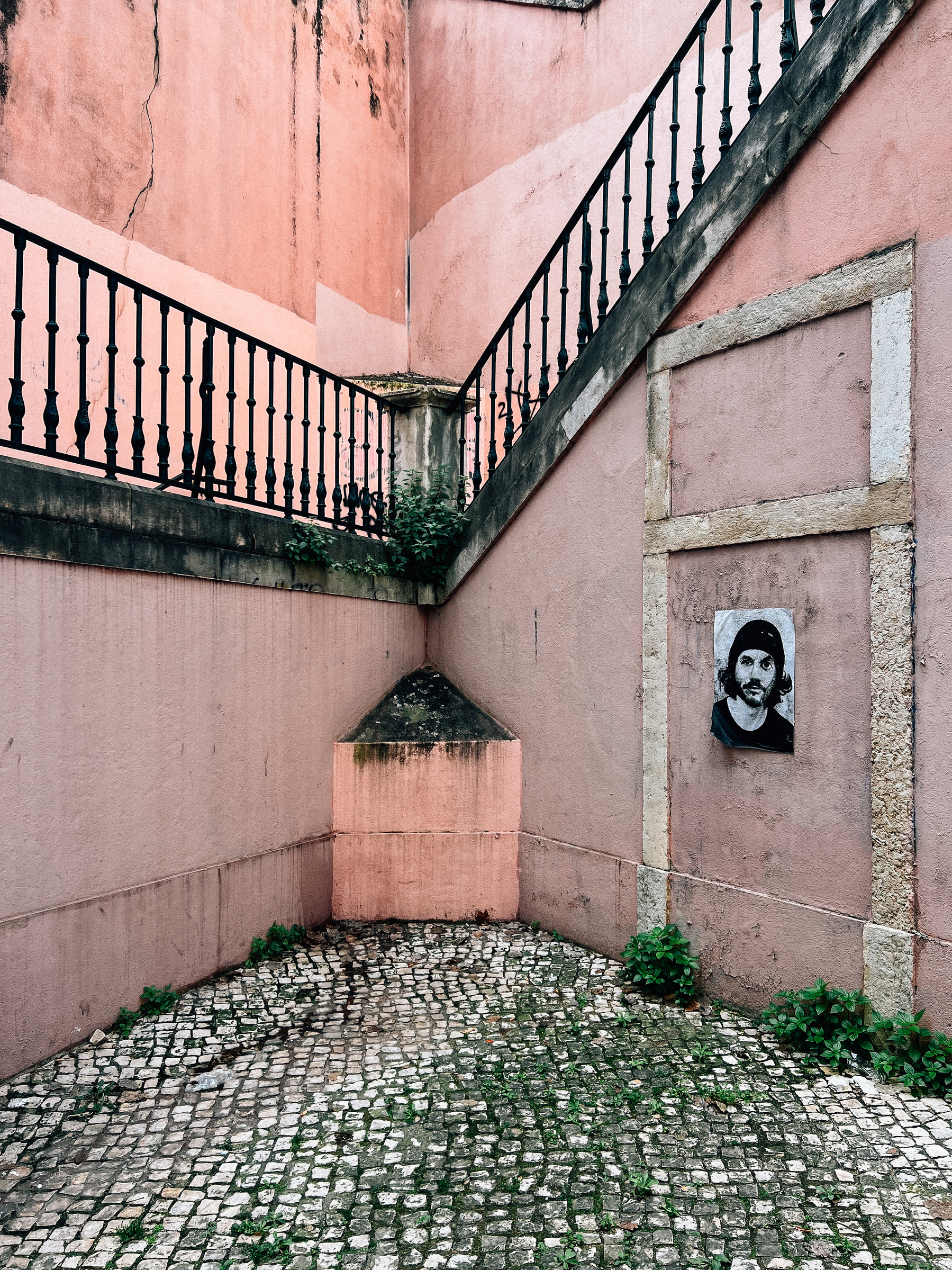 An outdoor corner of a building with weathered pink walls and a metal railing staircase. There&rsquo;s a black and white portrait posted on the wall and cobblestone pavement below.