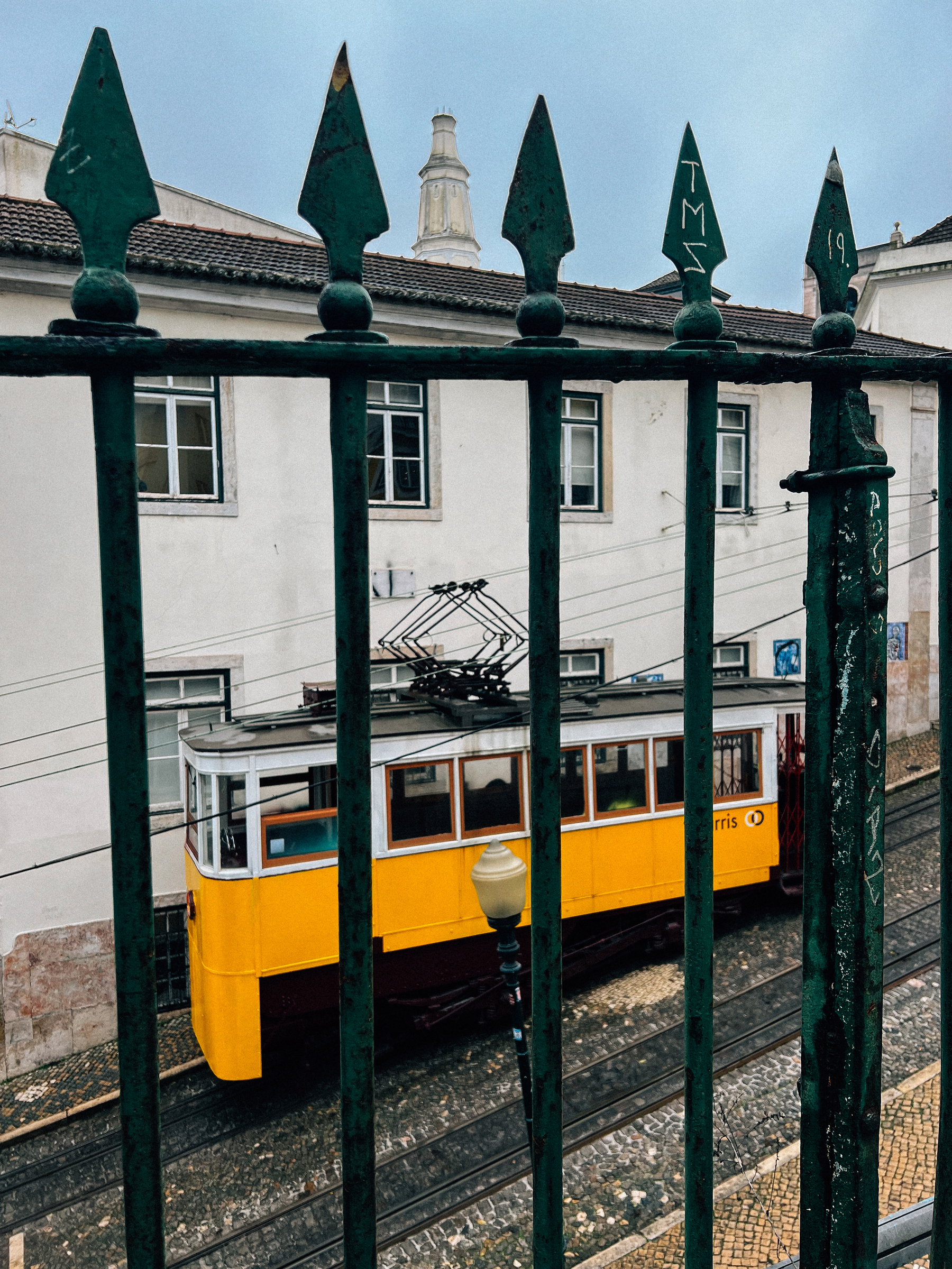 A classic yellow tram seen through a green wrought-iron fence, with traditional architecture and a lamppost in the background.