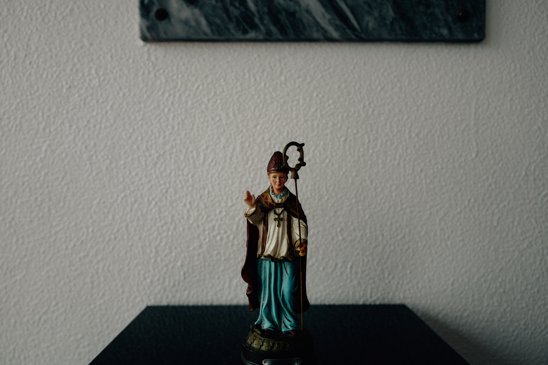 A figurine of a saintly figure in religious attire, likely representing a Catholic saint, holding a crosier and gesturing with one hand. The figure is set against a plain wall with a dark, abstract picture hanging above.