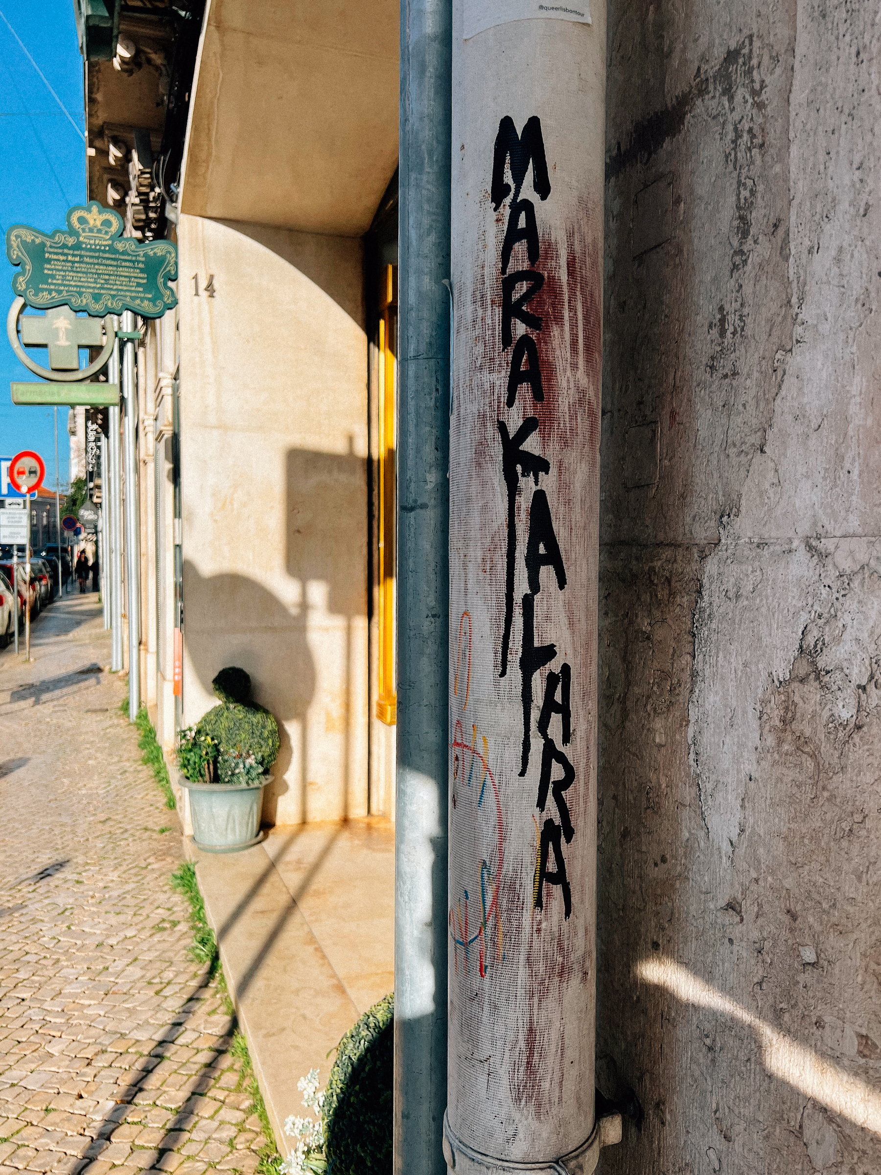 Graffiti on a column with the word &ldquo;MARAKALARA.&rdquo; Nearby is a street sign and potted plants along a cobblestone sidewalk, with a view down a quiet street.