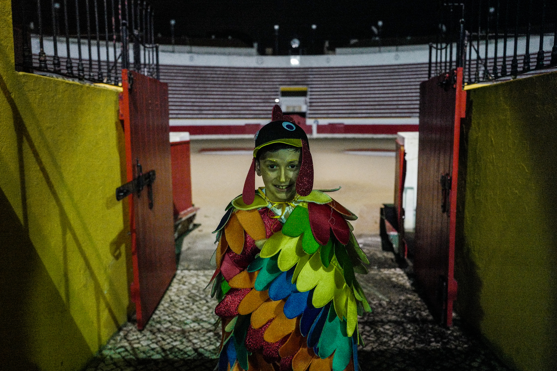 A child in a colorful bird costume stands at the entrance of a bullfighting arena at night.