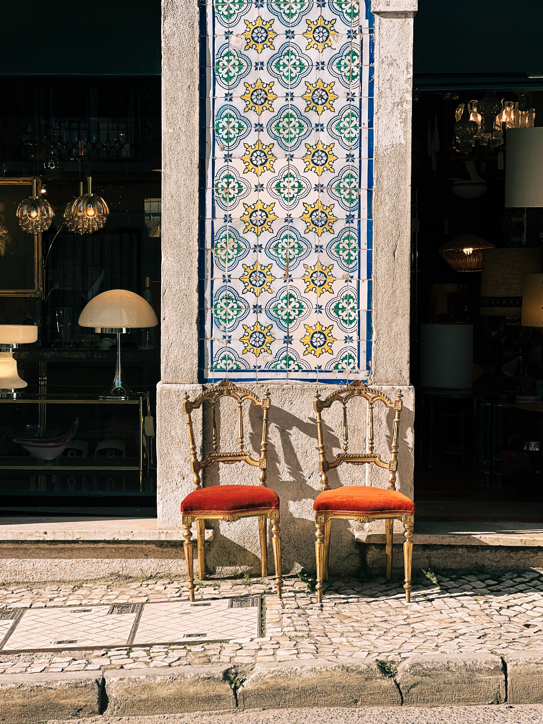 Two vintage red chairs set against a tiled wall featuring a colorful pattern of ceramic tiles, with a glimpse of a stylish interior through a window to the left.