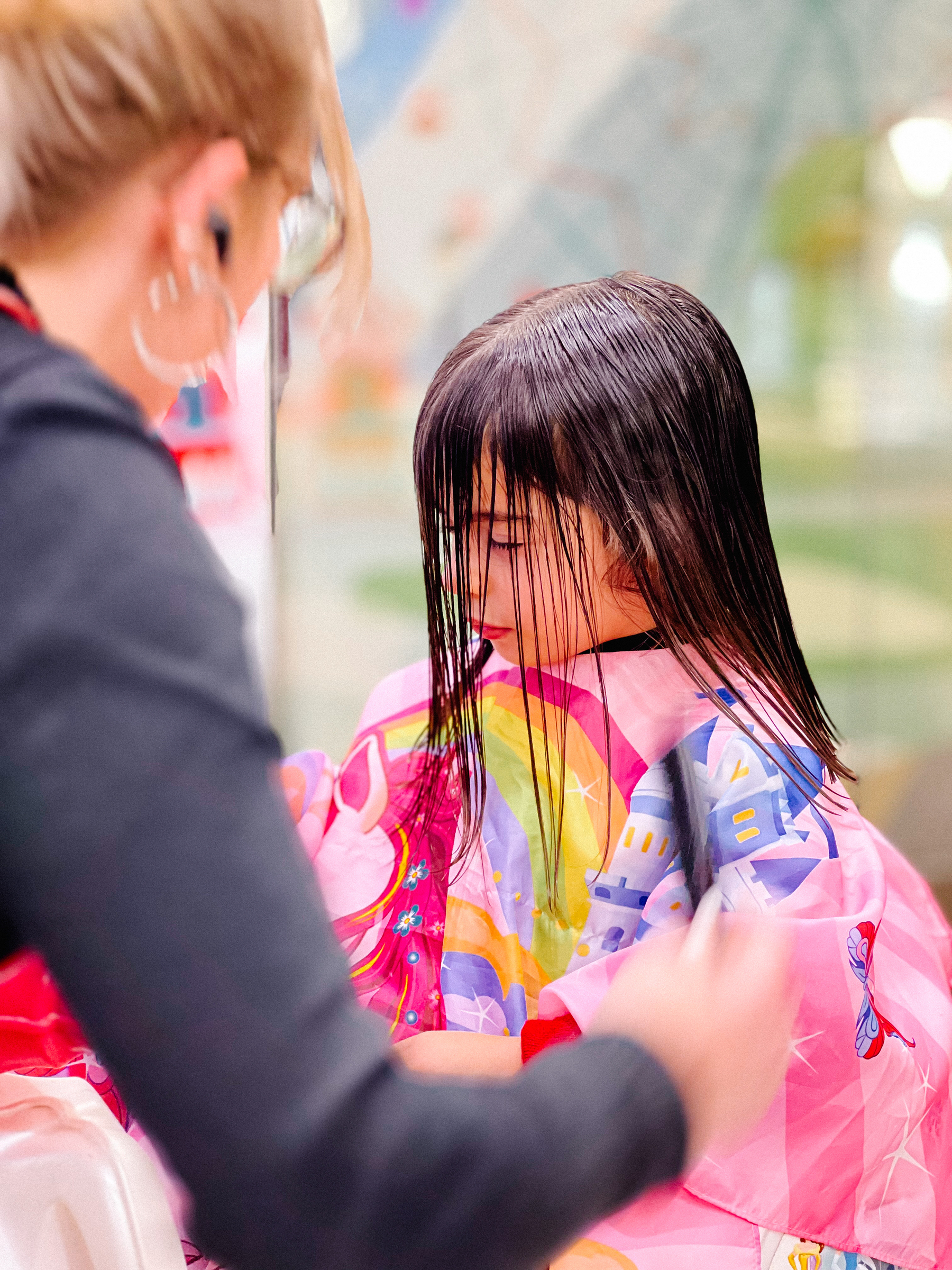 A young child having a haircut, with wet hair covering their face, while a hairstylist works on their hair. The child is wearing a colorful cape with cartoon designs.