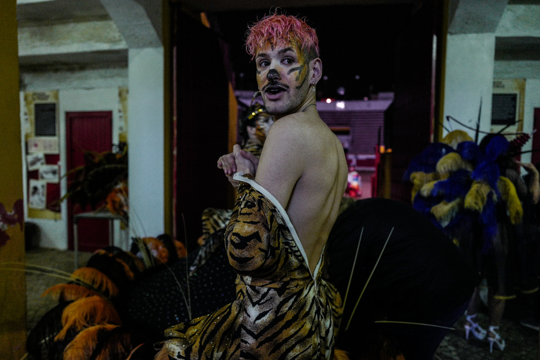 A person with pink hair and tiger face paint is looking back over their shoulder while wearing a striped tiger costume with a tail. There are props and costumes in the blurry background, suggesting a theatrical or festive setting.