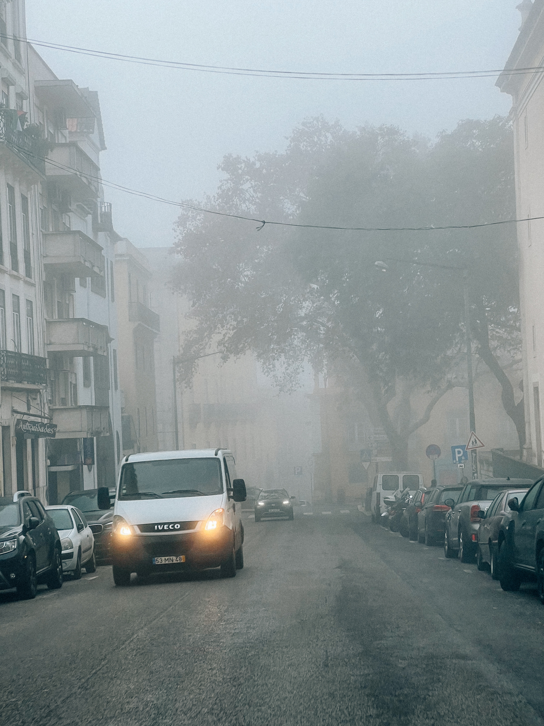 A foggy street with parked cars lining both sides, a white IVECO van with its headlights on is in the foreground driving towards the camera, and faint outlines of buildings and trees are visible in the background.