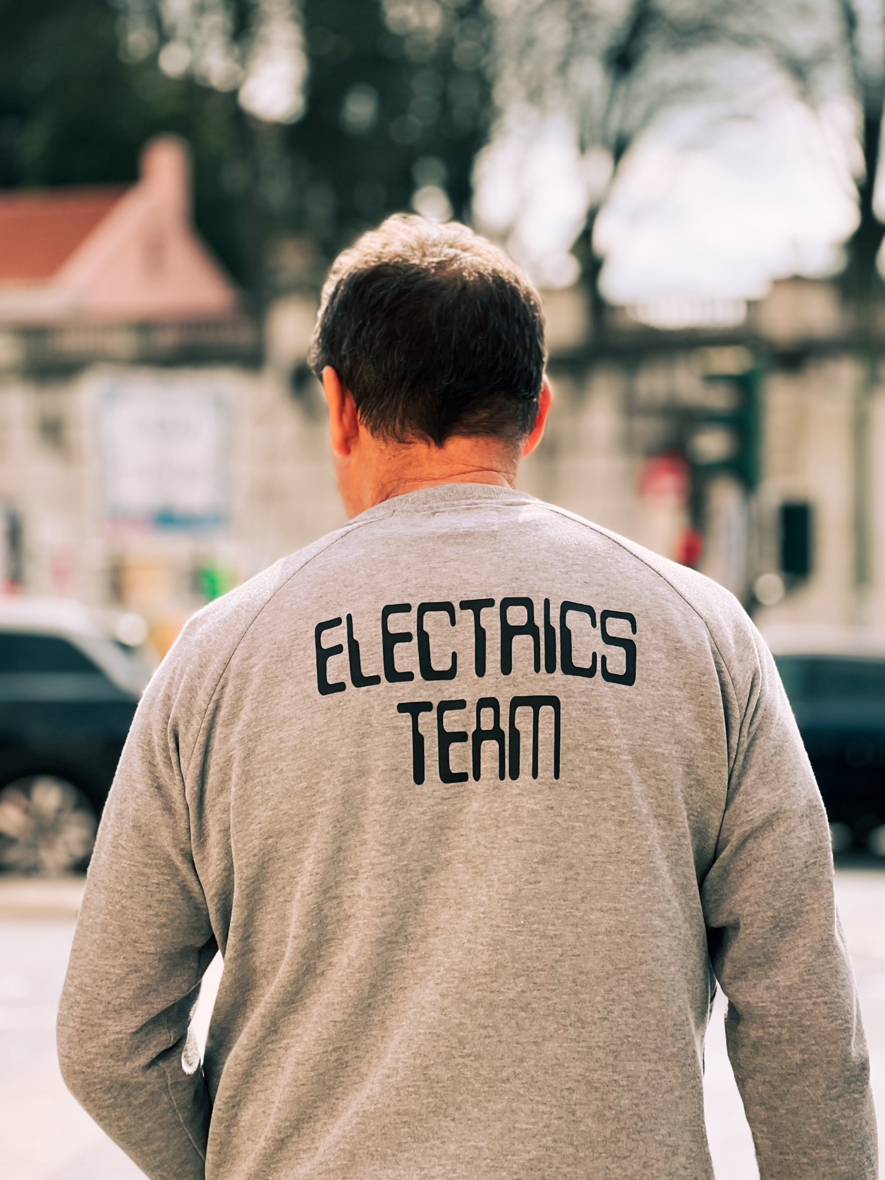 A man seen from behind wearing a grey shirt with the words &ldquo;ELECTRICS TEAM&rdquo; on the back. The background is slightly blurred showing an urban setting with trees, a road, and vehicles.