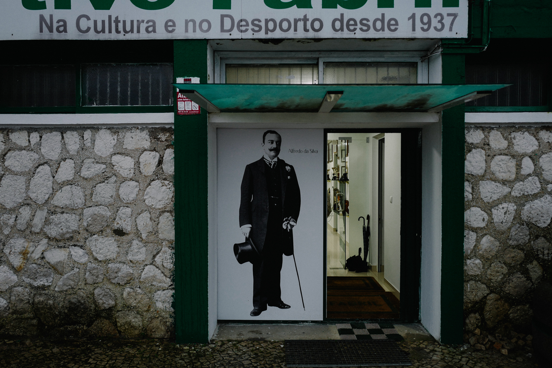 An entrance to a building with stone walls and an overhanging sign reading &ldquo;CULTURA Na Cultura e no Desporto desde 1937.&rdquo; A black and white photograph of a man in a suit and top hat is displayed in the doorway.