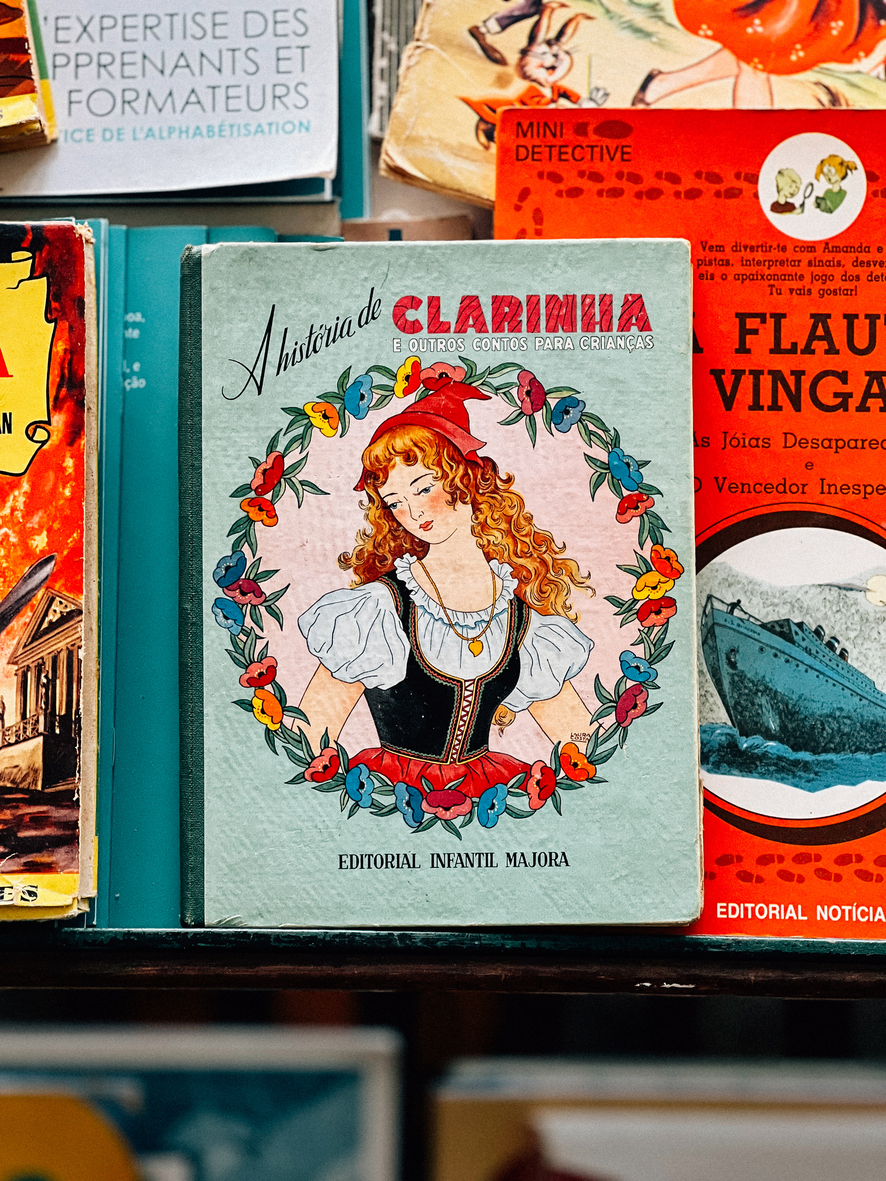 A vintage book titled &ldquo;A Historia de Clarinha e outros contos para crianças&rdquo; on a bookshelf amidst other old books. The cover features an illustration of a young girl with red hair wearing a renaissance-style red hat and dress.