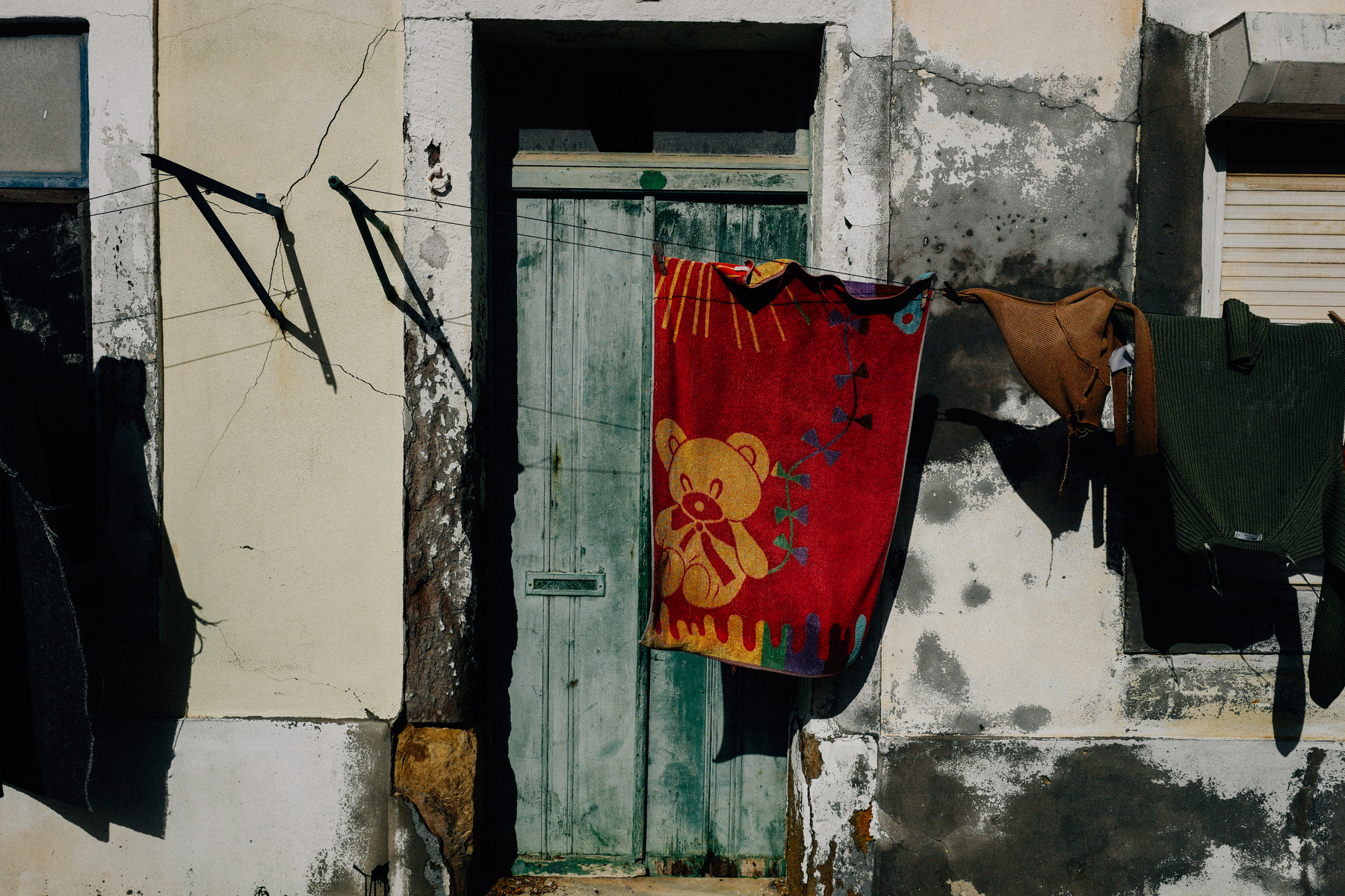 An old weathered wall with a green door, above which hangs a window with broken shutters. Laundry, including a red towel with a teddy bear design and several dark colored clothes, is hung out to dry on a makeshift clothesline.