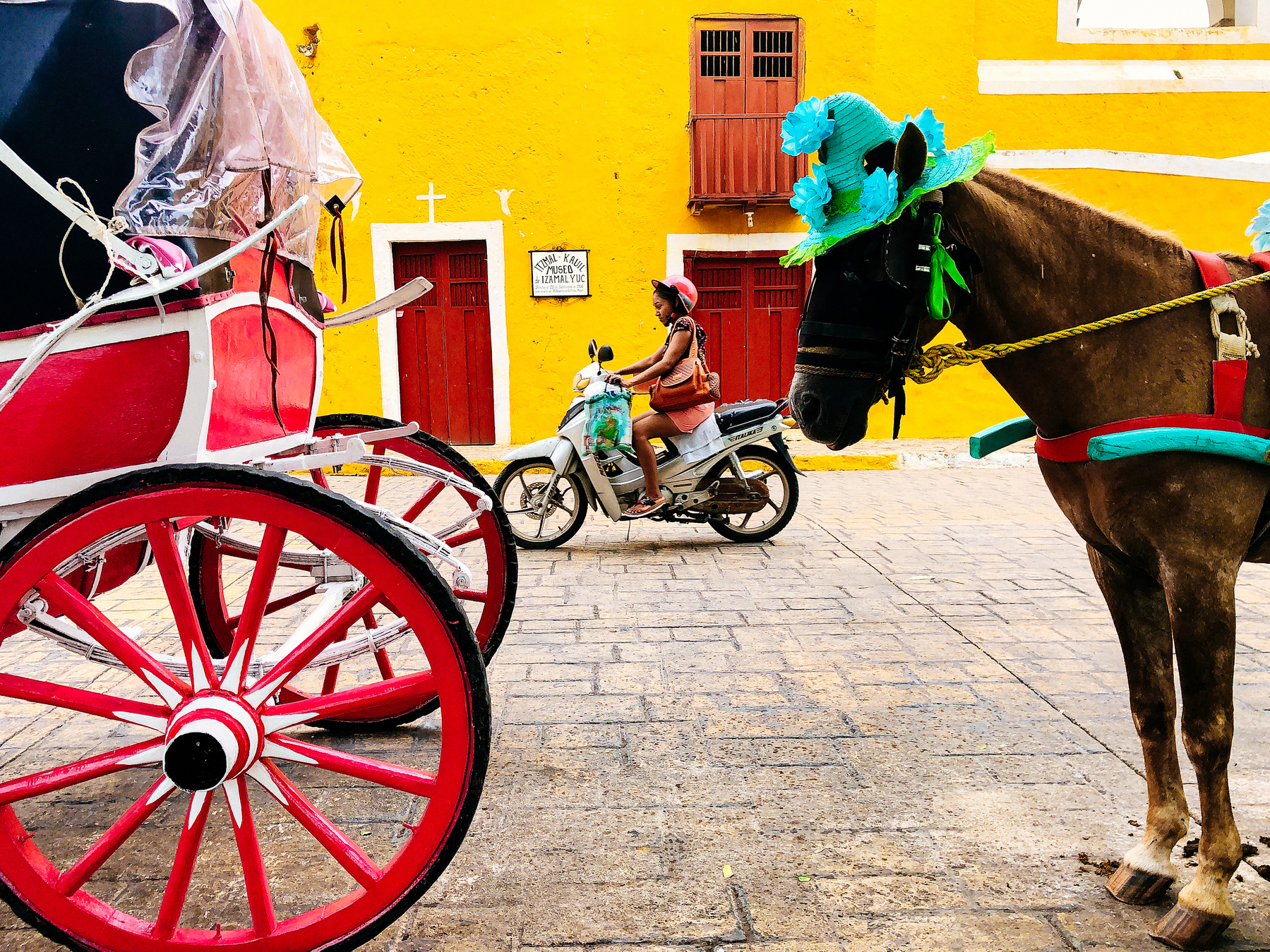 A horse with blue adornments, and a red and white carriage, stand on a paved street next to a building with a yellow facade and a red door. In the background, a person rides a scooter past another red door. 