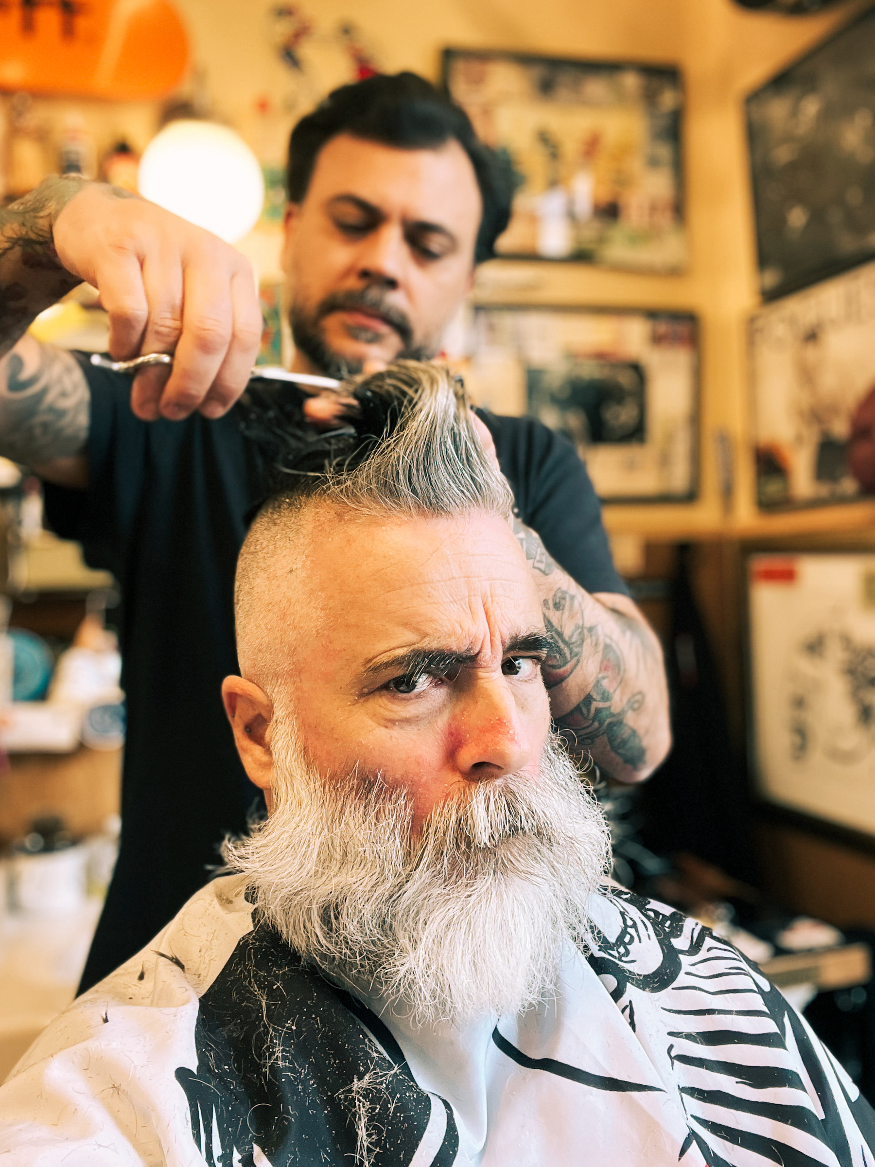 A barber with tattoos is styling a client&rsquo;s hair in a barber shop. The client, with a prominent white beard, is seated and looking intensely at the camera. The scene has a vintage feel with decor on the walls. 